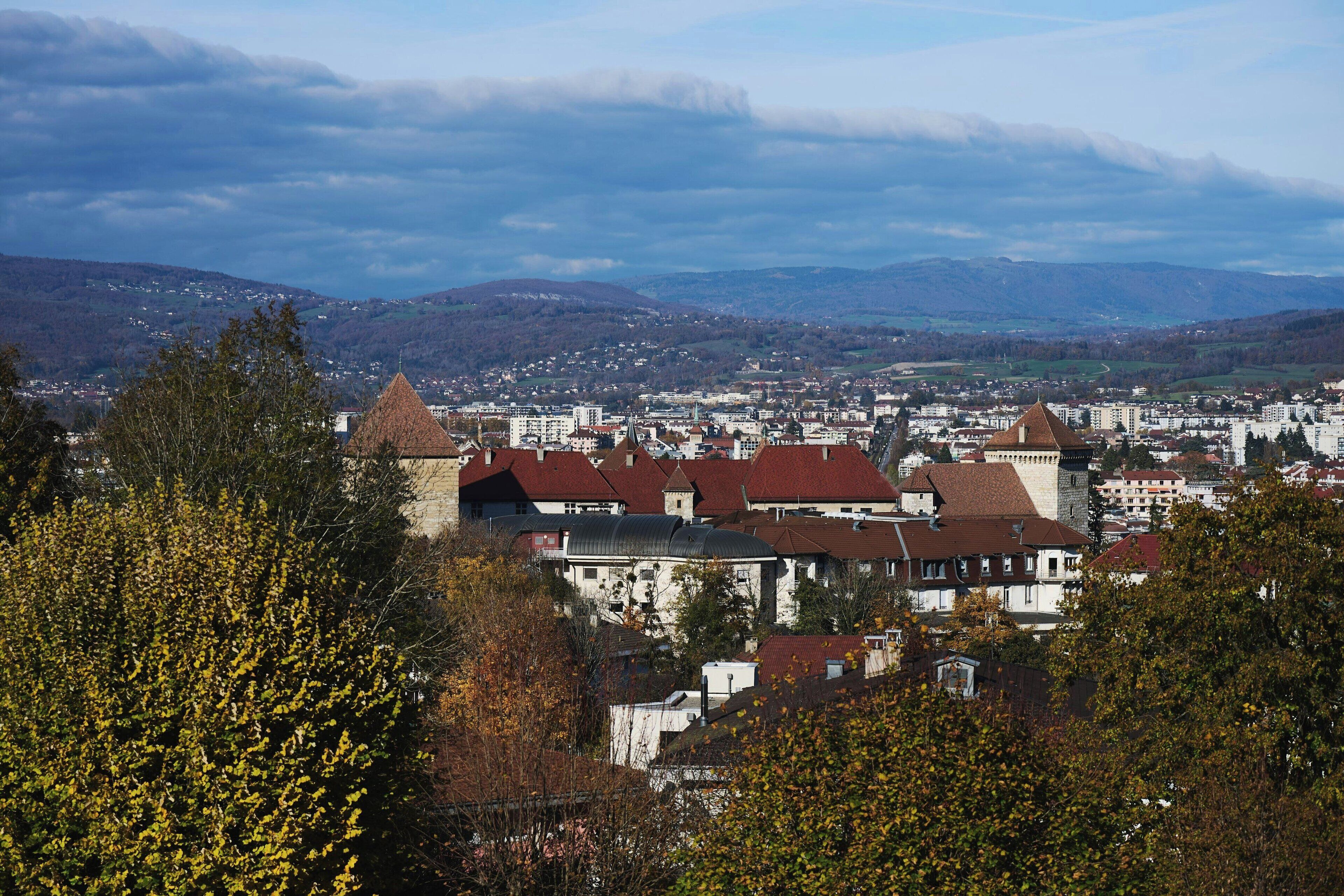 A town with historic buildings, including a castle-like structure with a conical roof, surrounded by modern buildings and a mountainous backdrop.