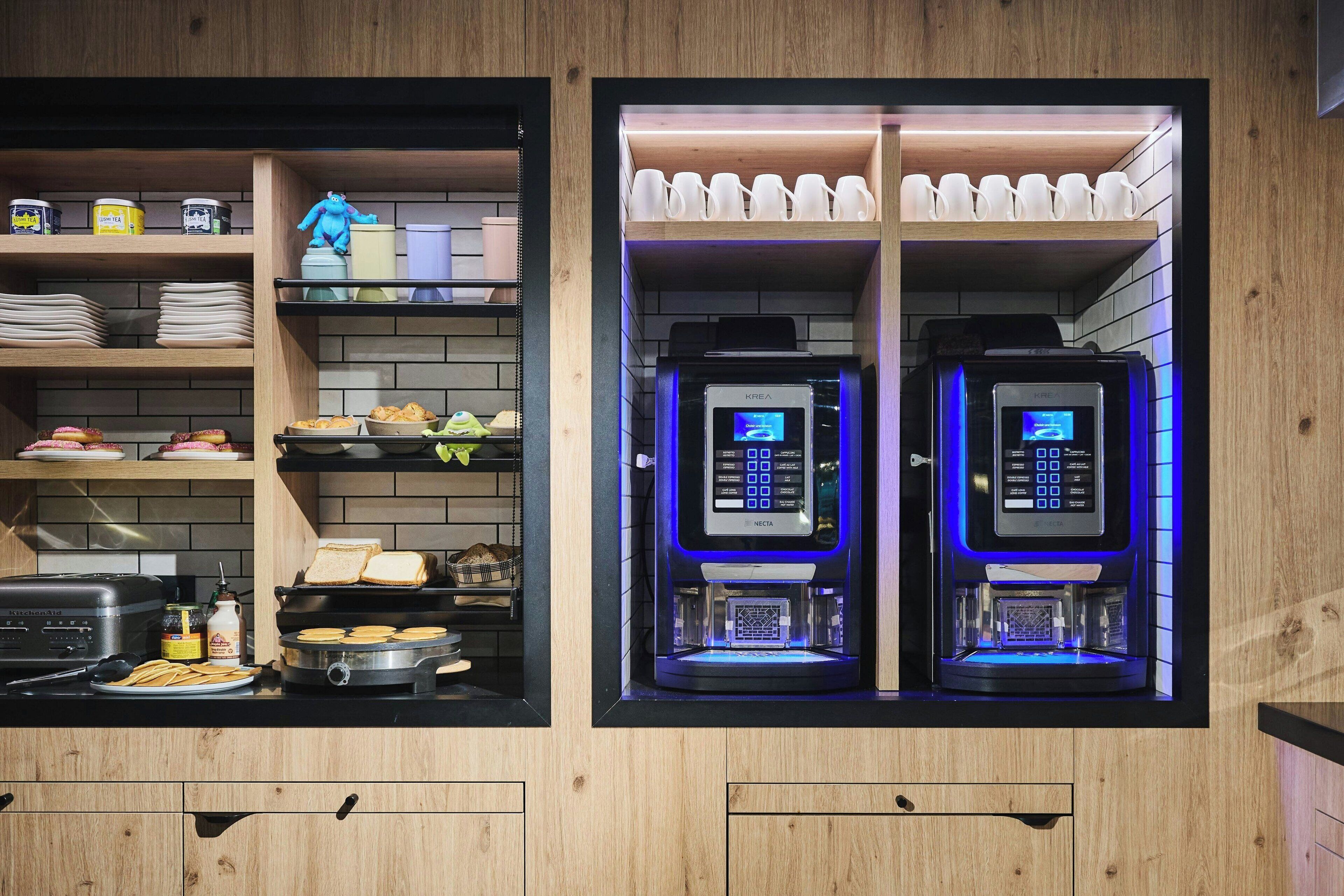 A kitchen with two coffee machines, a toaster, and various food items on shelves.