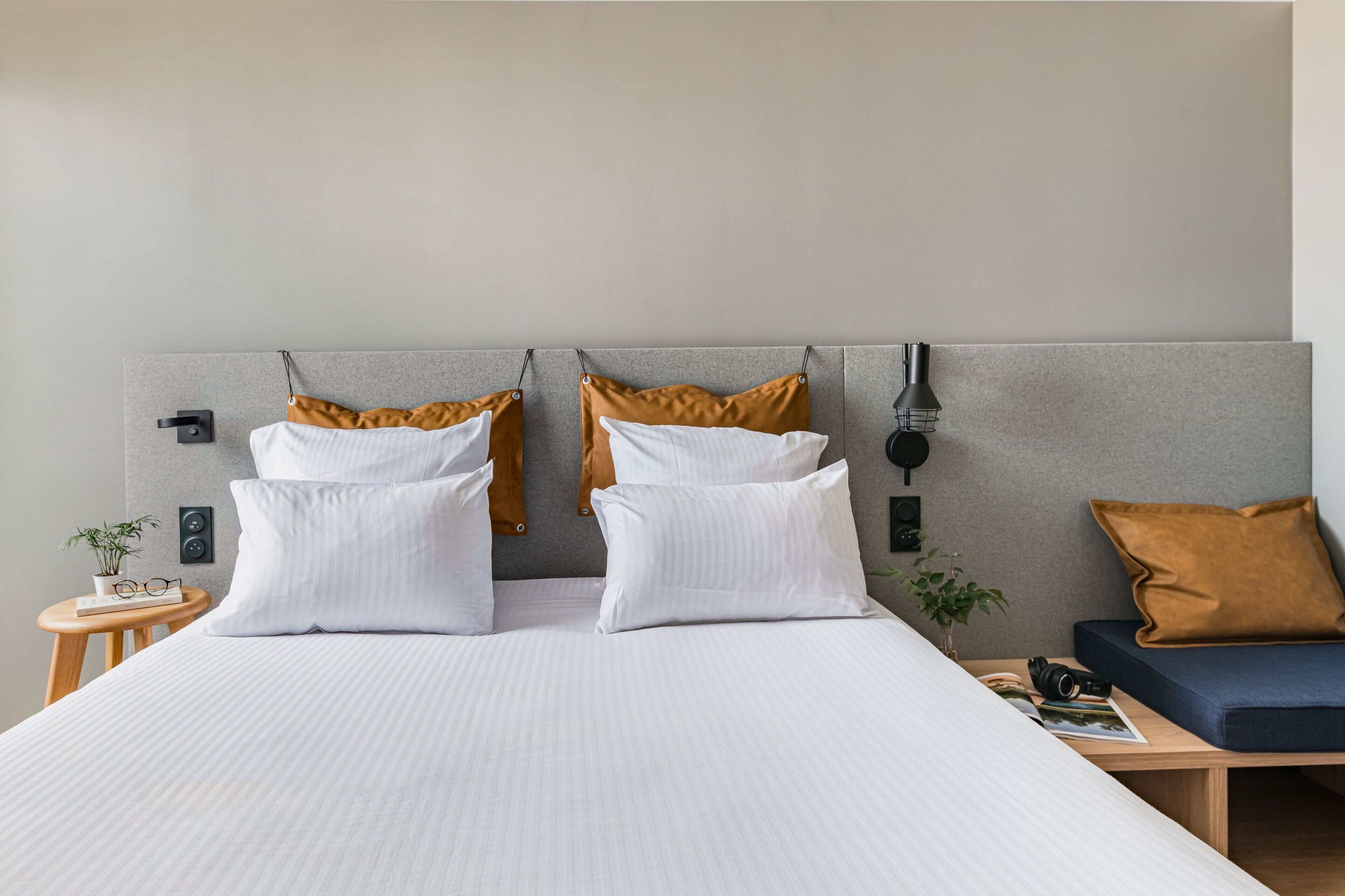 A neatly made bed with white linens and two pillows, a bedside table with a plant, and a small seating area with a cushion and a lamp.