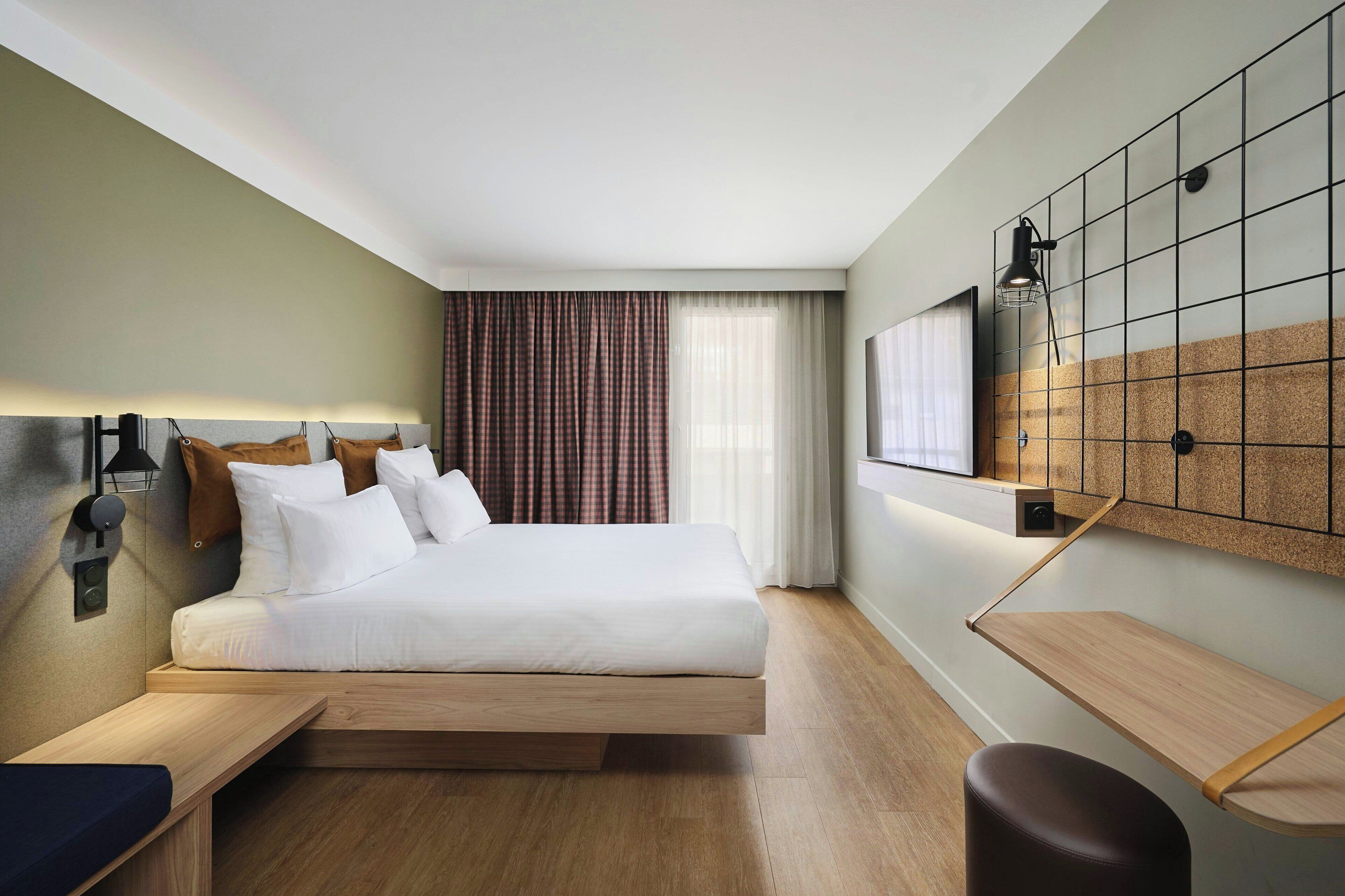 A modern hotel room with a large bed, bedside tables, a desk, and a corkboard wall.
