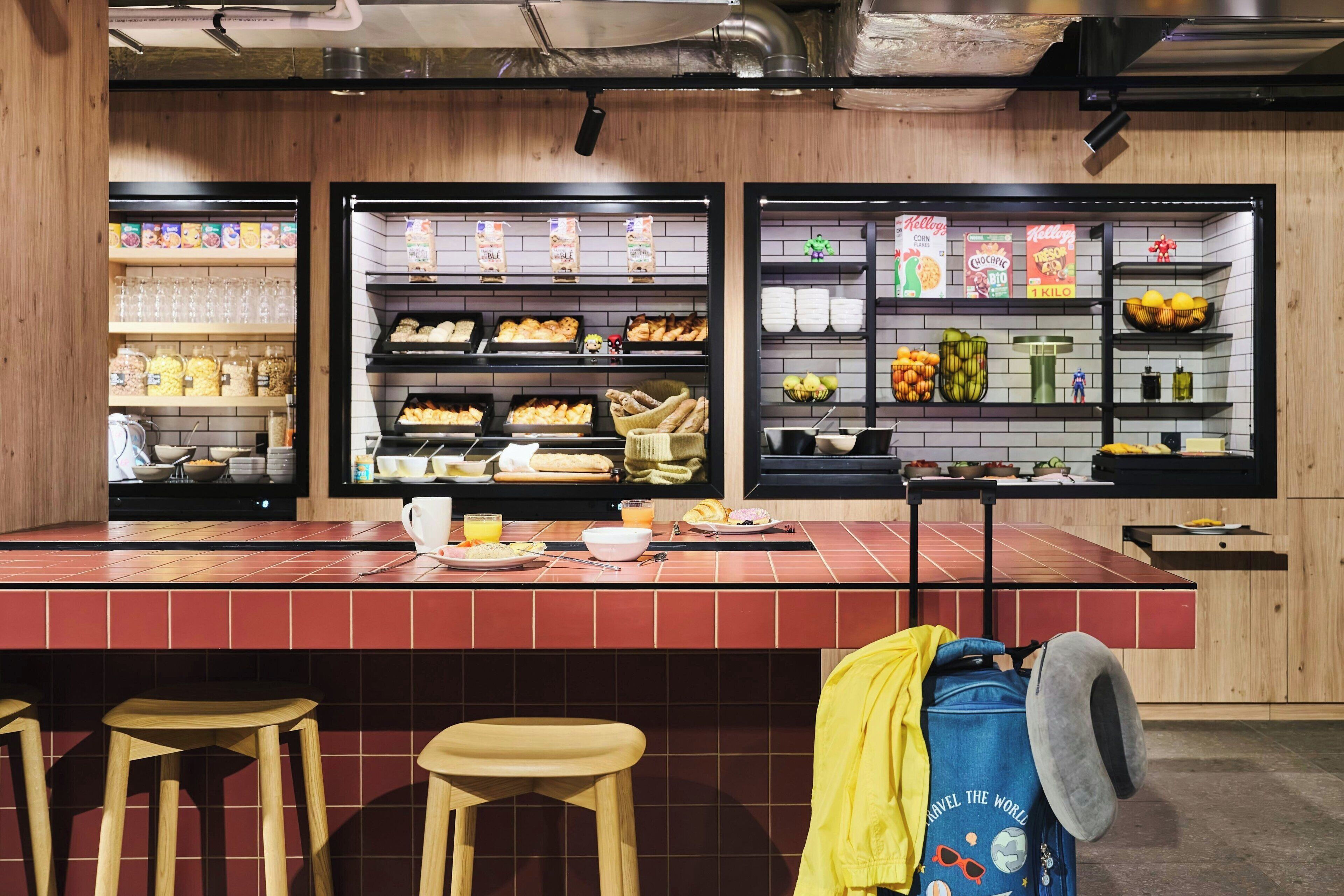 Un bar pour le petit-déjeuner, avec des étagères remplies de céréales, de pain et de fruits. On y trouve des tabourets et une table où sont disposés les aliments pour le petit-déjeuner.