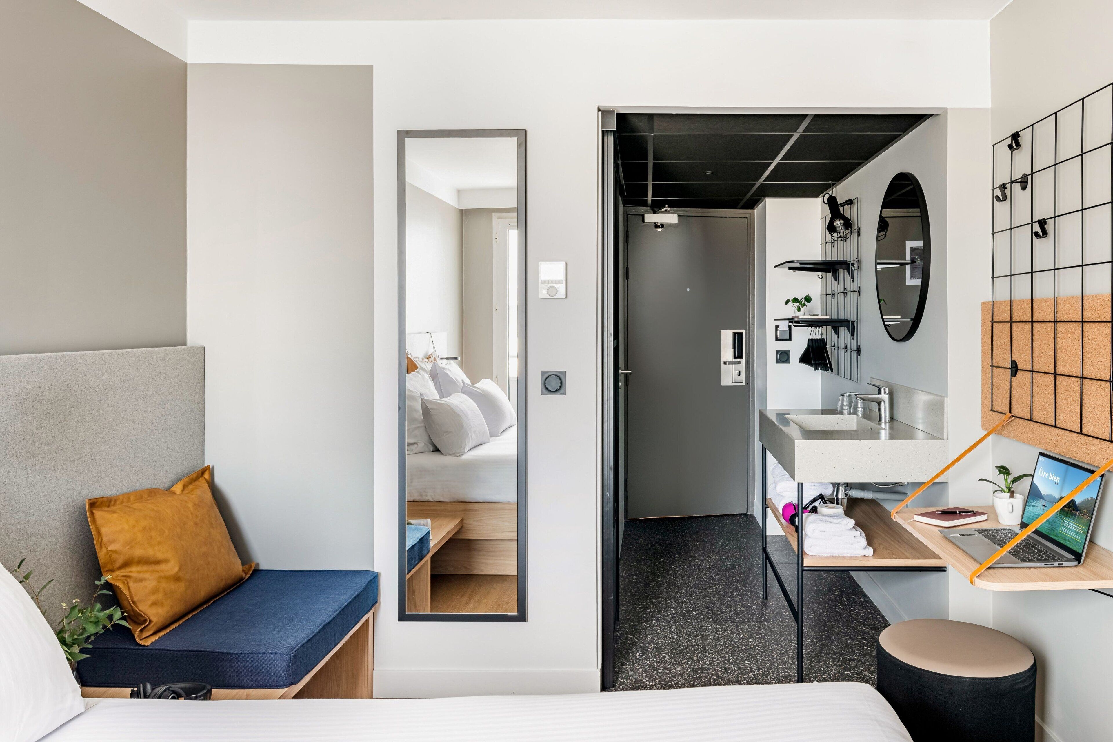 A modern hotel room with a bed, a small desk with a laptop, a mirror, and a view into the bathroom.