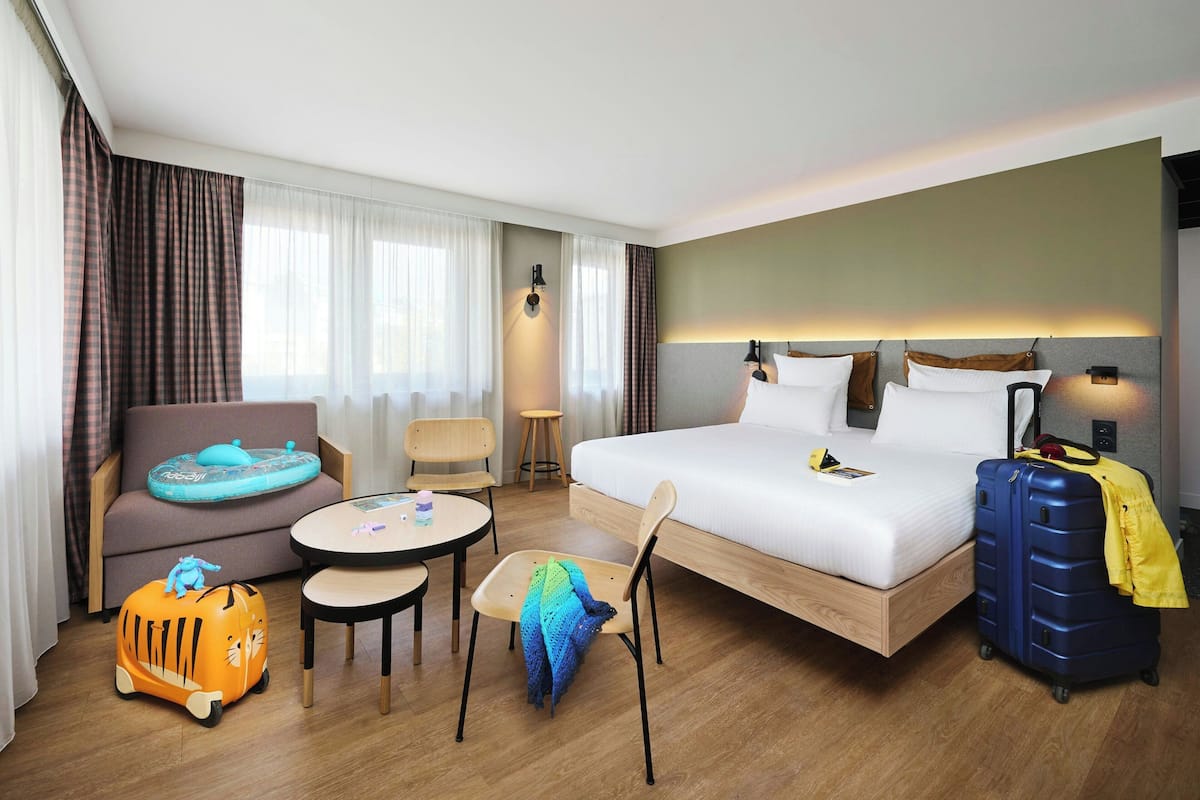 Une chambre d’hôtel avec un lit, un canapé, une petite table, une chaise, une valise et une lampe.