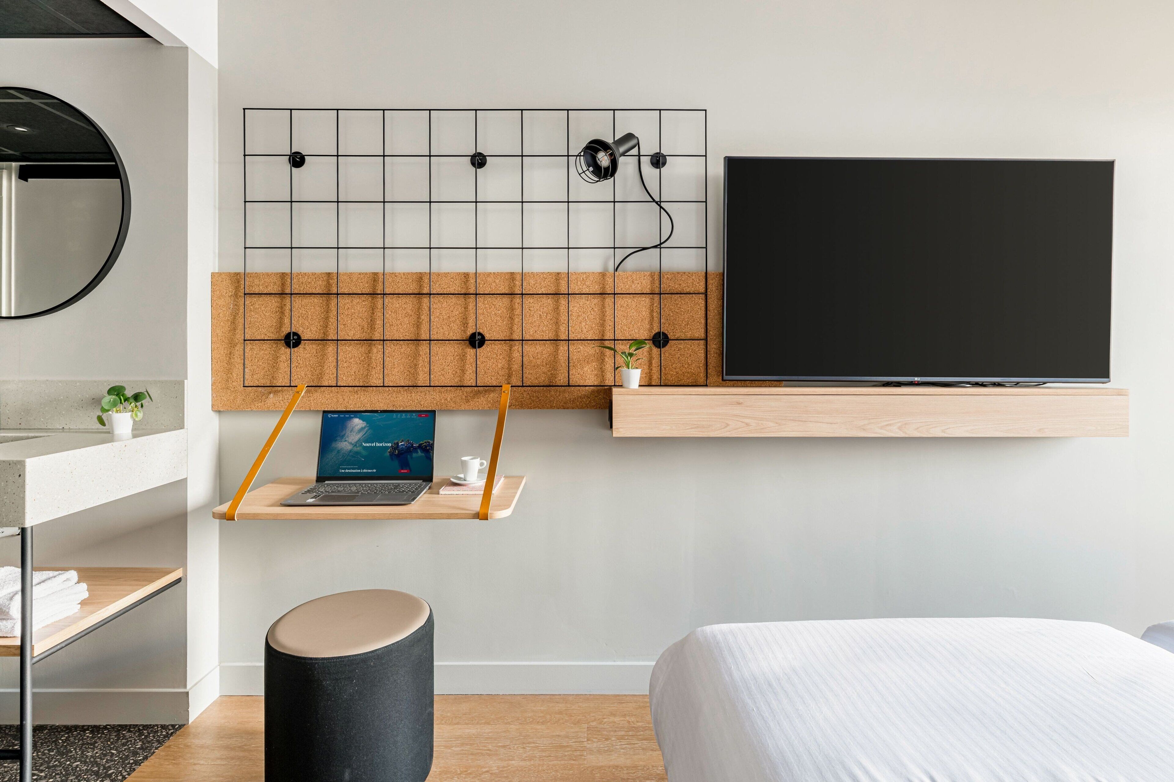 A modern hotel room with a large bed, a mounted TV, a desk with a laptop, a small plant, and a round stool.