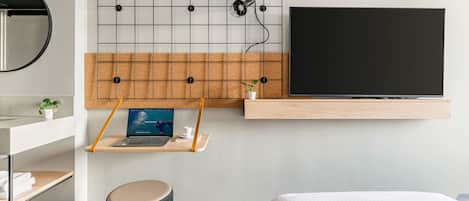 A modern hotel room with a large bed, a mounted TV, a desk with a laptop, a small plant, and a round stool.