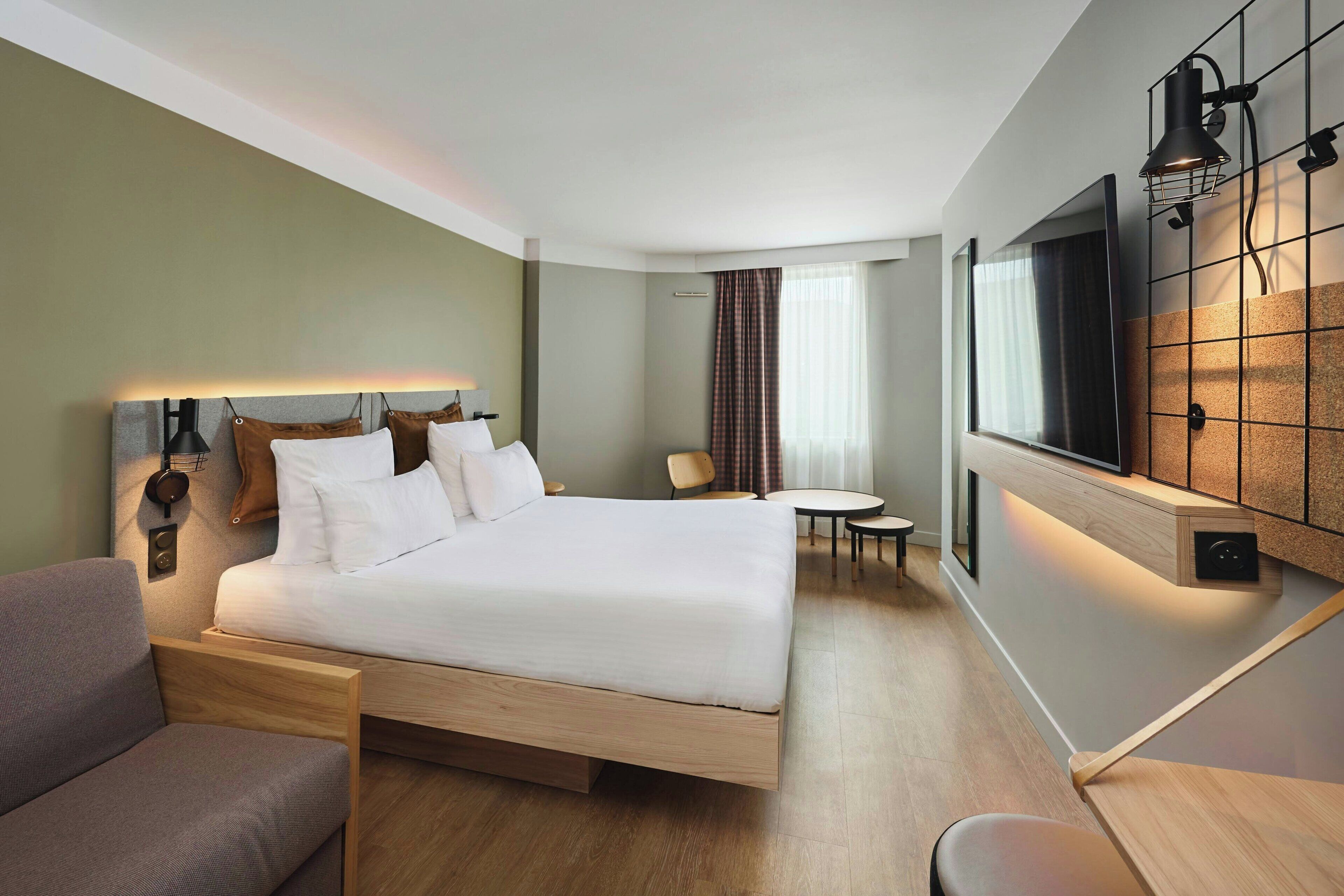 A modern hotel room with a large bed, a TV mounted on the wall, a small round table, and a sofa.