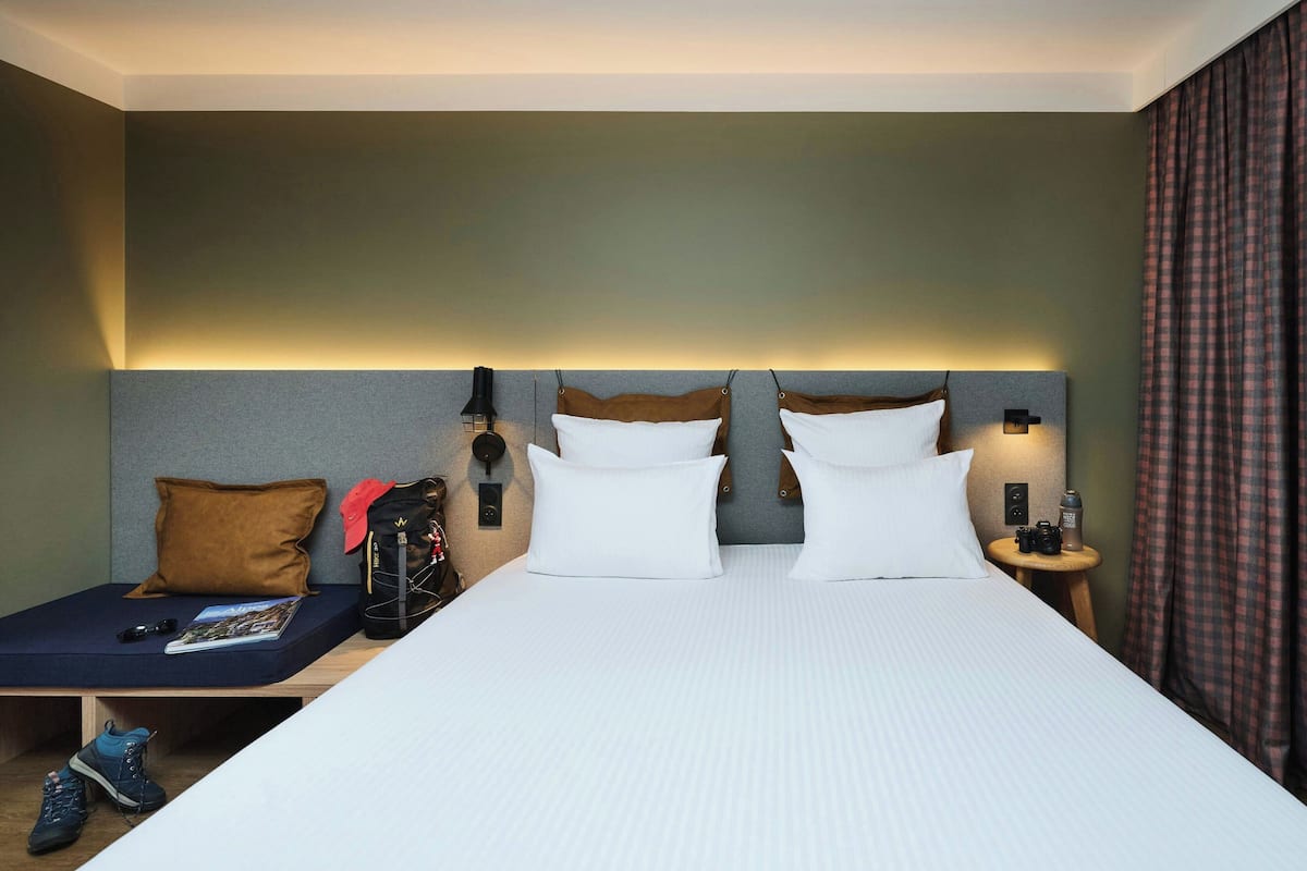 Une chambre d’hôtel avec un grand lit, une table de chevet, une commode, une lampe fixée au mur et une fenêtre avec des rideaux.
