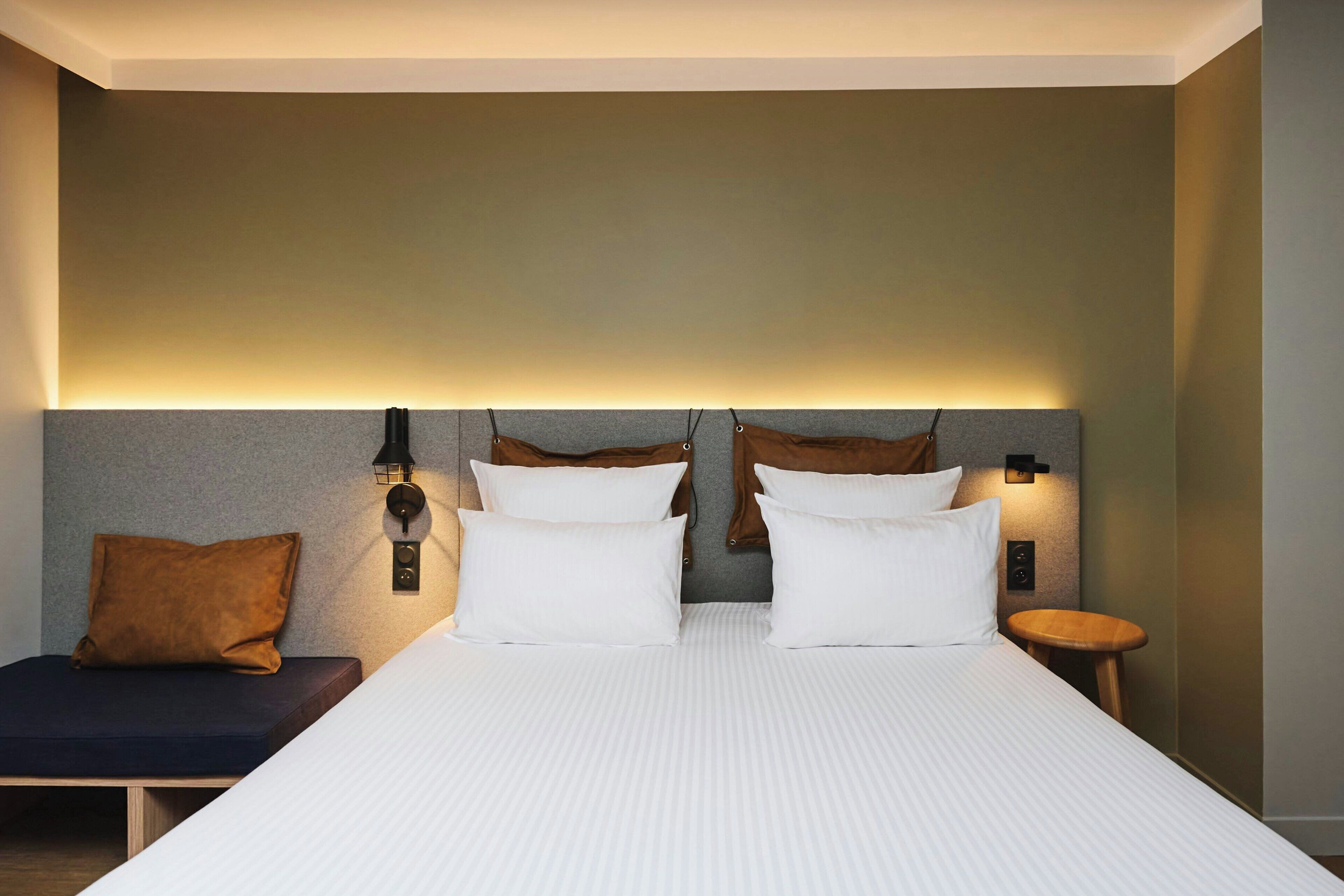A modern hotel room with a large bed, two bedside tables, a nightstand, and a bench.