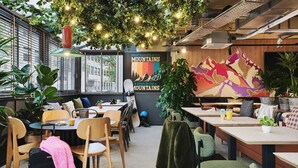 A cozy café with wooden tables and chairs, a mural of mountains, and hanging plants. - Moxy Annecy (Annecy)