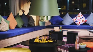 A cozy lounge area with a blue sofa, colorful cushions, a black coffee table with a fire feature, and a large floor lamp. - Moxy Annecy (Annecy)