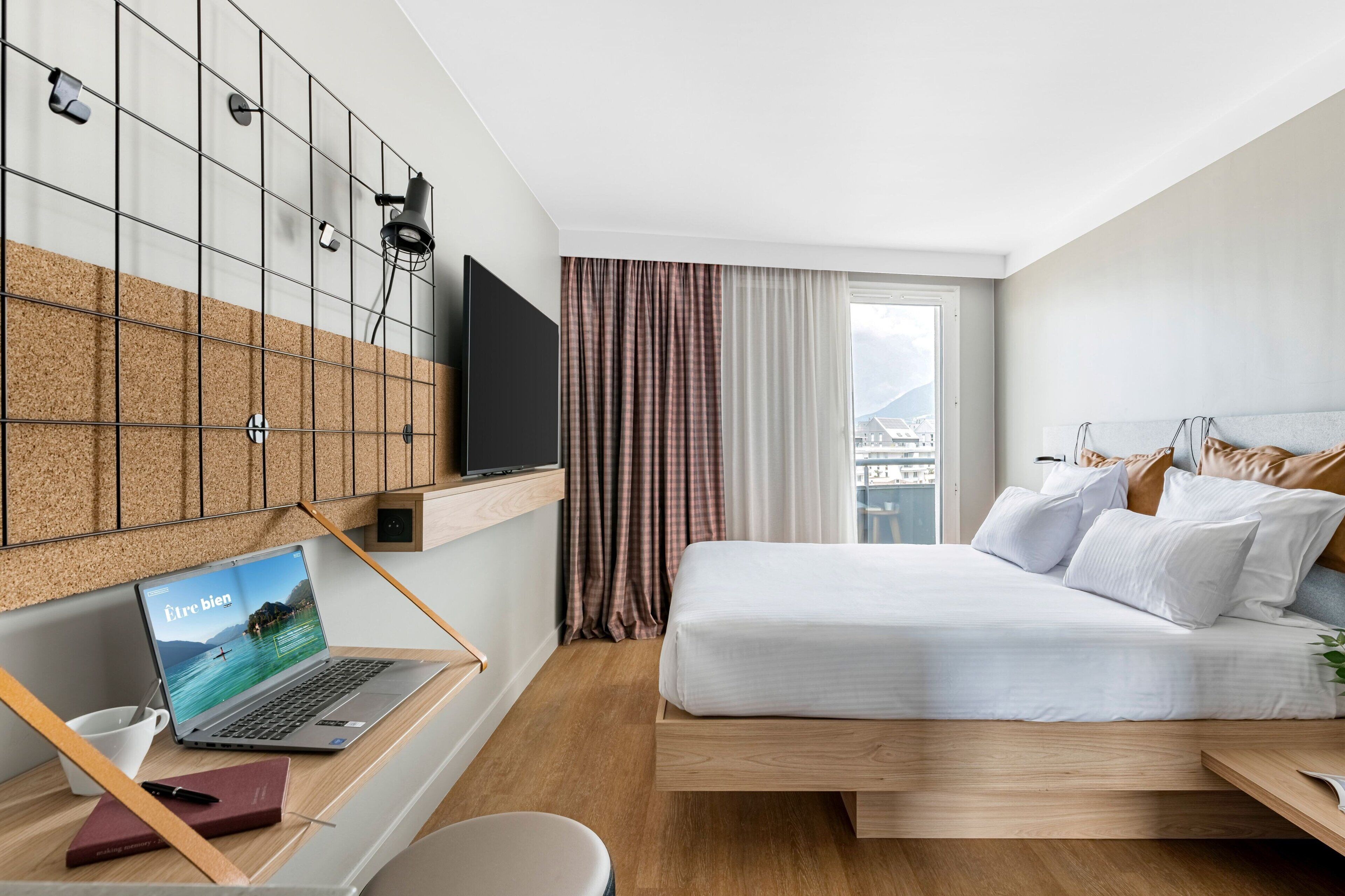 A modern hotel room with a bed, a desk with a laptop, a TV, and a view of the city.