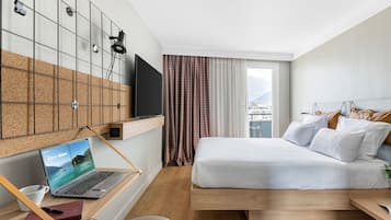 A modern hotel room with a bed, a desk with a laptop, a TV, and a view of the city.