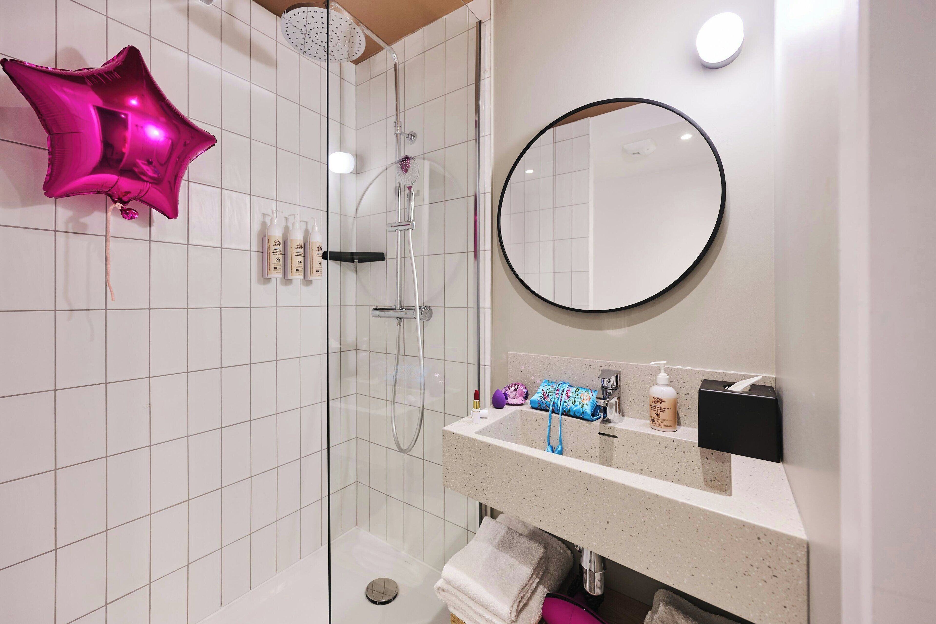 A modern bathroom with a glass-enclosed shower, a round mirror, and a vanity with toiletries.