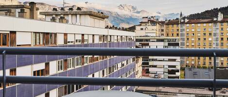 Un balcón con una mesa y sillas, con vista a un paisaje urbano que incluye edificios y montañas al fondo.