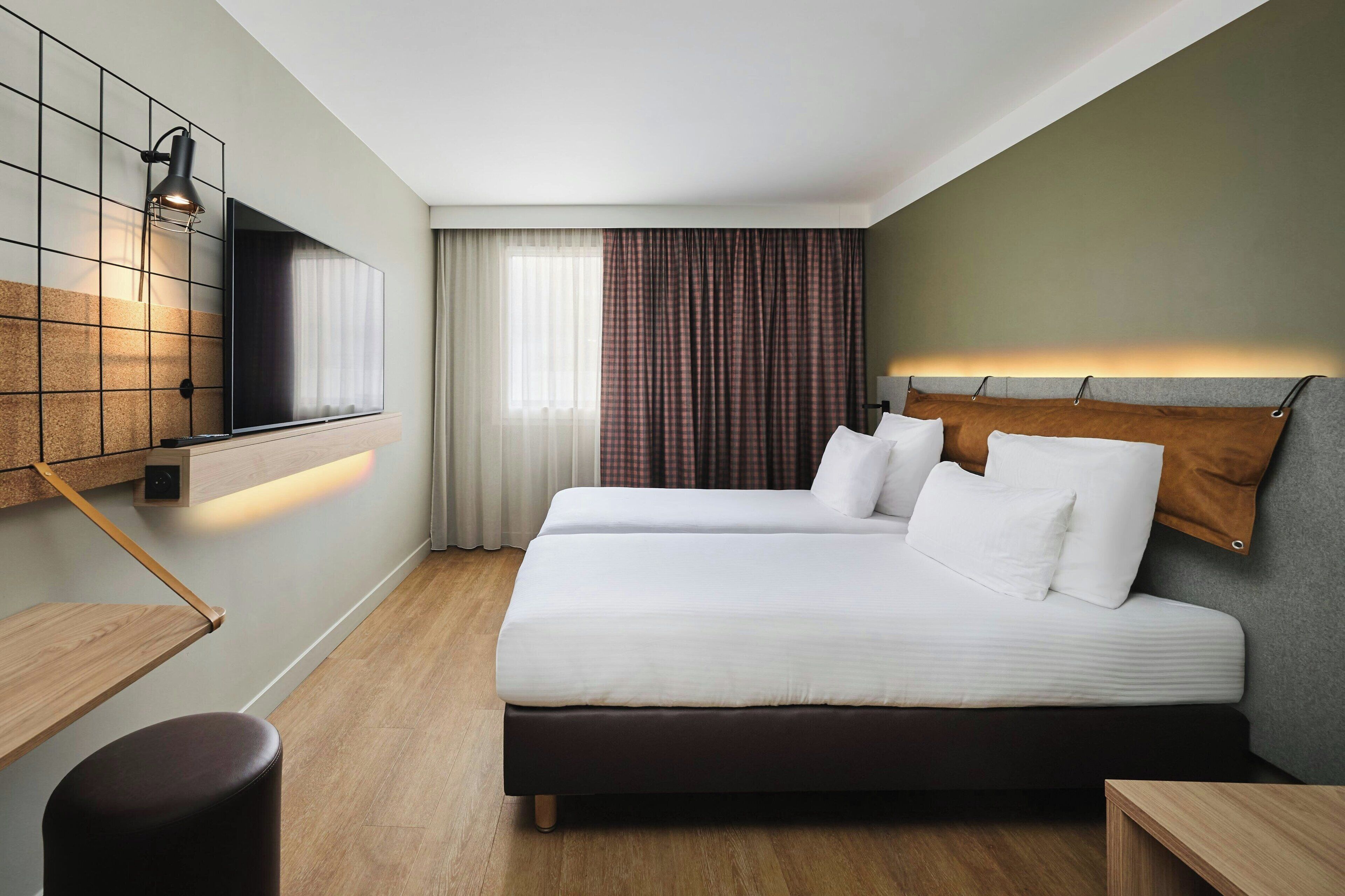 A hotel room with a large bed, a TV mounted on the wall, a wooden floor, and a window with curtains.