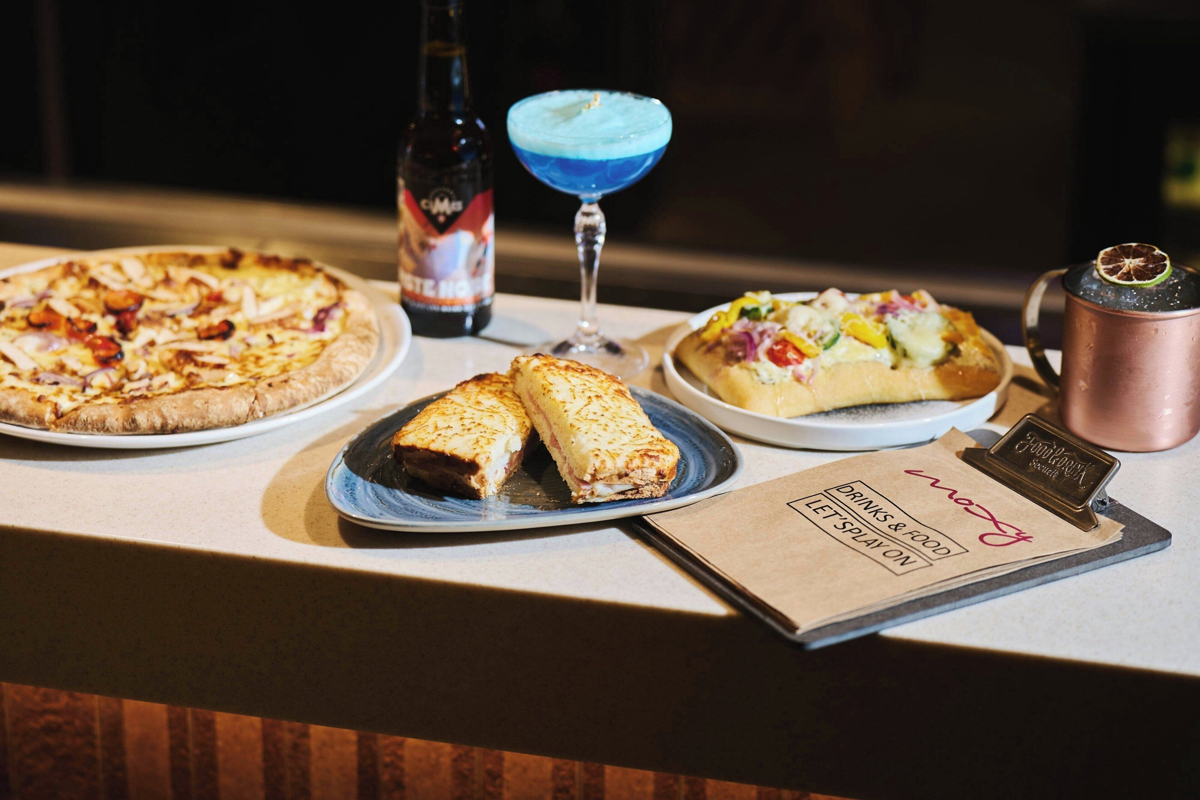 A bar area with a variety of food items including pizzas and a dessert, a bottle of beer, and a blue cocktail.