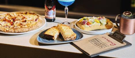 A bar area with a variety of food items including pizzas and a dessert, a bottle of beer, and a blue cocktail.