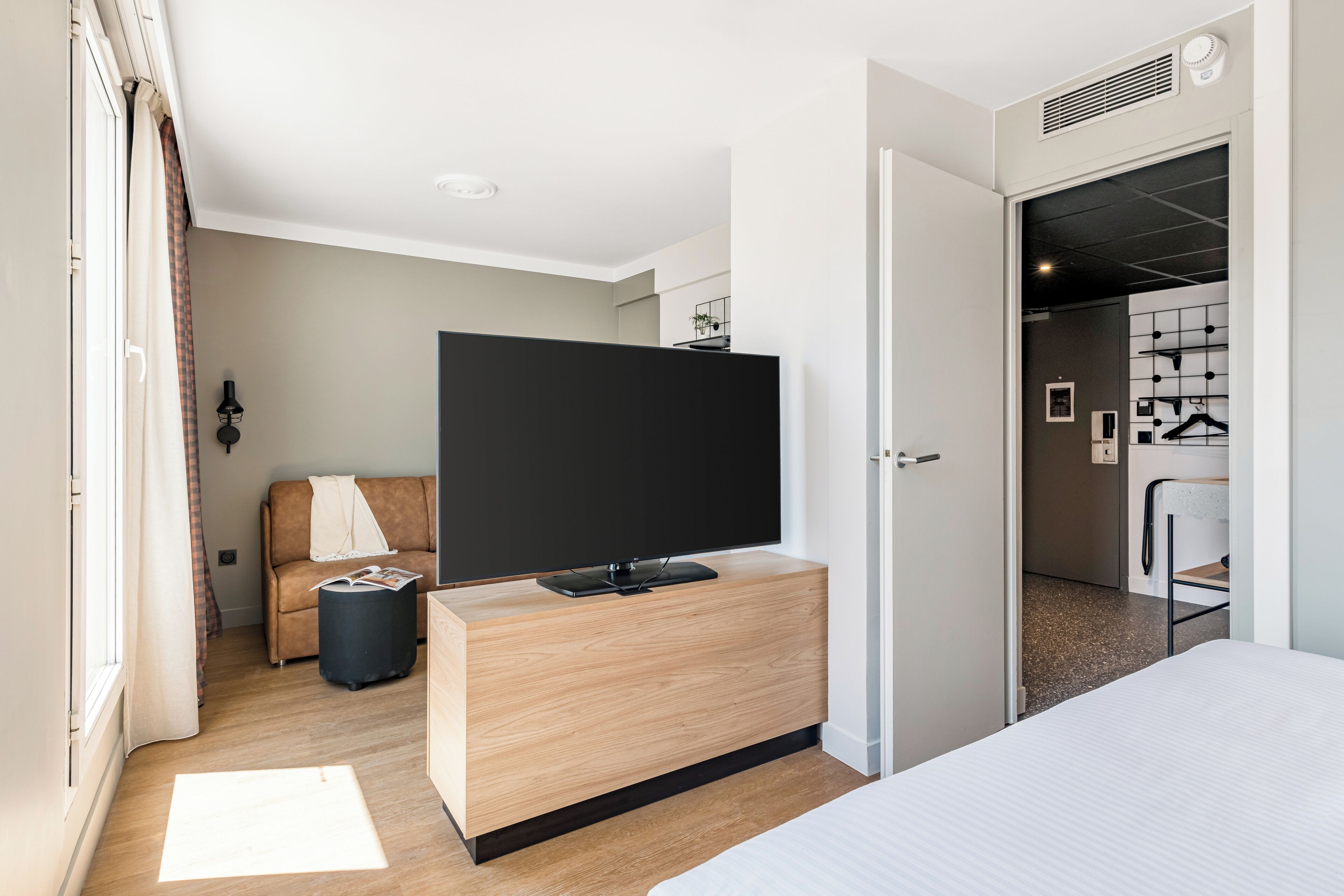 Habitación de hotel moderna con un televisor de pantalla plana grande sobre una consola de madera, un sofá, una mesita auxiliar y una cama.