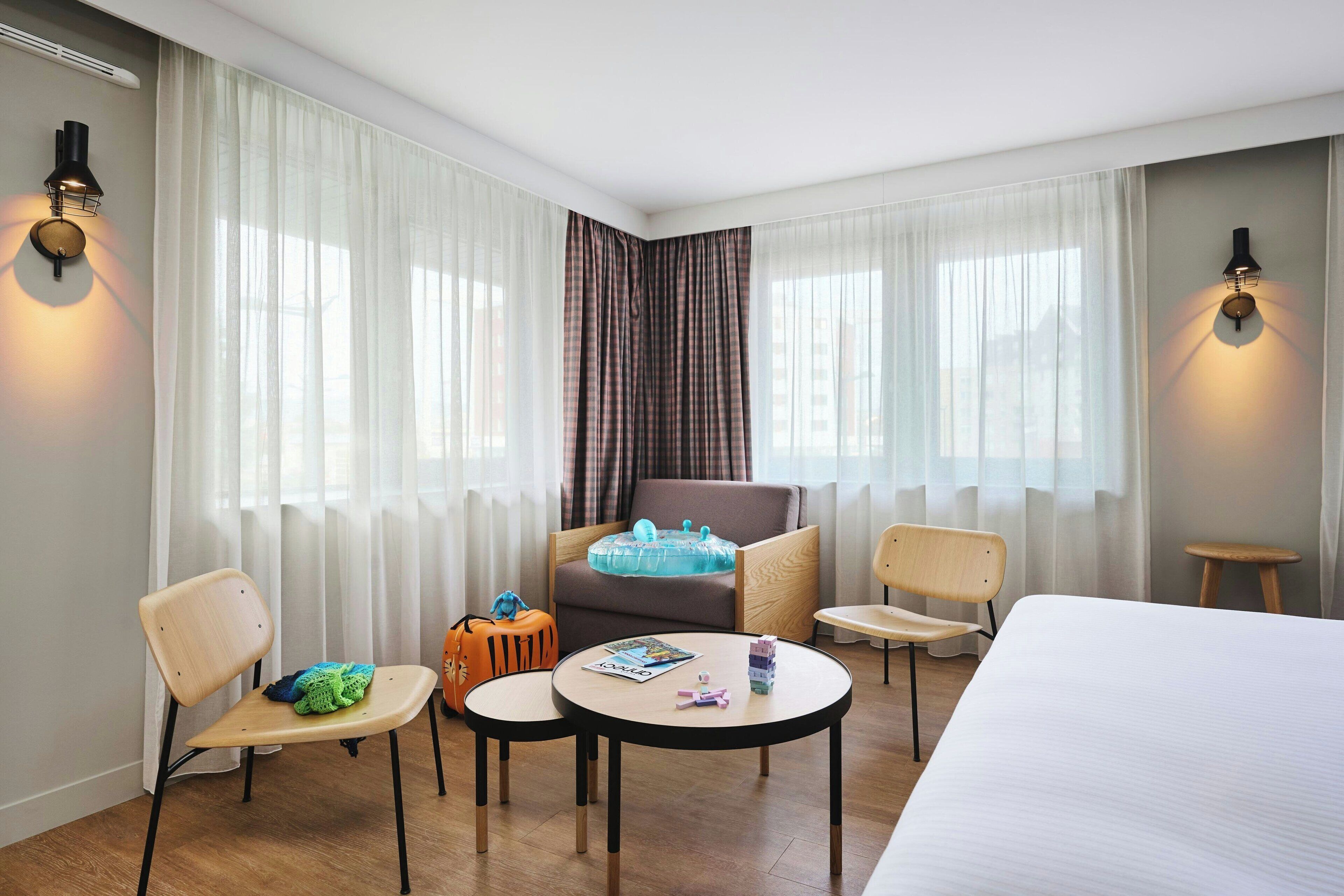 A hotel room with a bed, two chairs, a small table, a sofa, and a window with sheer curtains.