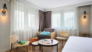 A hotel room with a bed, two chairs, a small table, a sofa, and a window with sheer curtains.