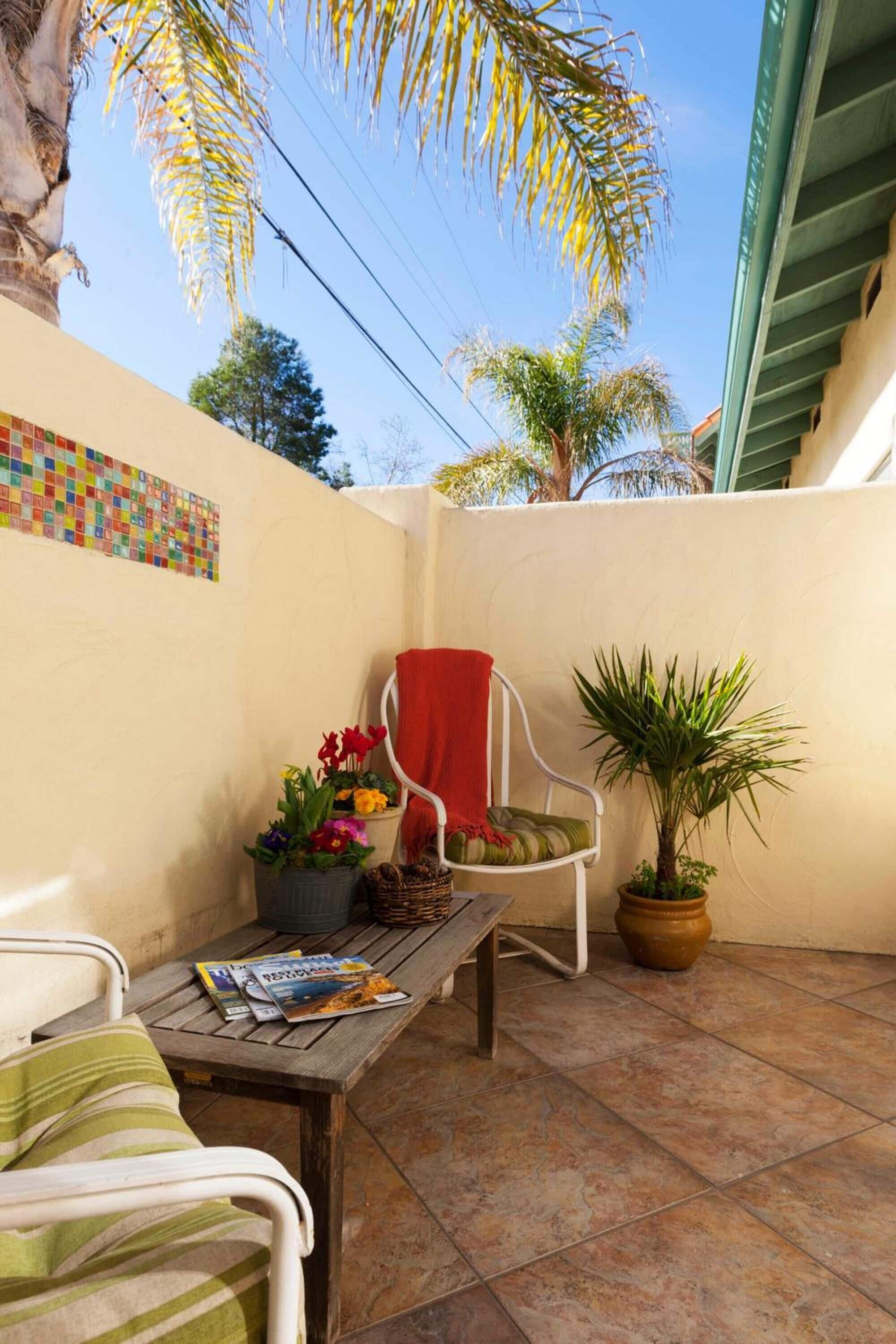 Un pequeño patio con una silla, una mesa con revistas, una planta en maceta y una pared con un mosaico colorido.