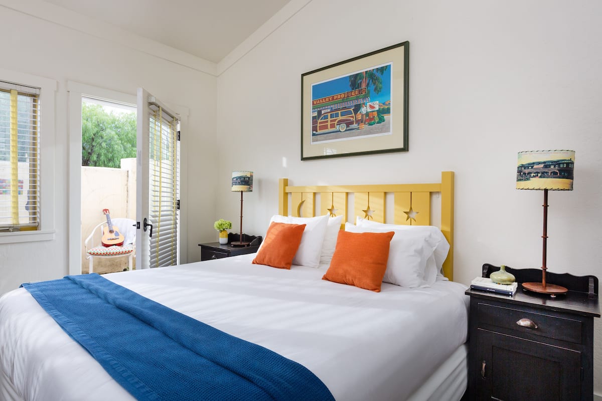 A neatly made bed with white and orange pillows, a blue throw blanket, and a framed picture of a vintage car on the wall.