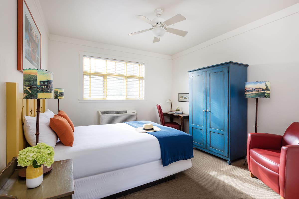 A hotel room with a bed, a desk, a chair, a wardrobe, and a ceiling fan.