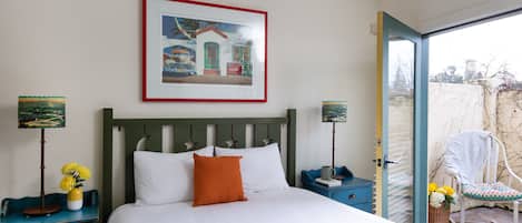 A neatly made bed with white linens and an orange pillow, a framed picture above it, and a blue nightstand with a vase of yellow flowers.