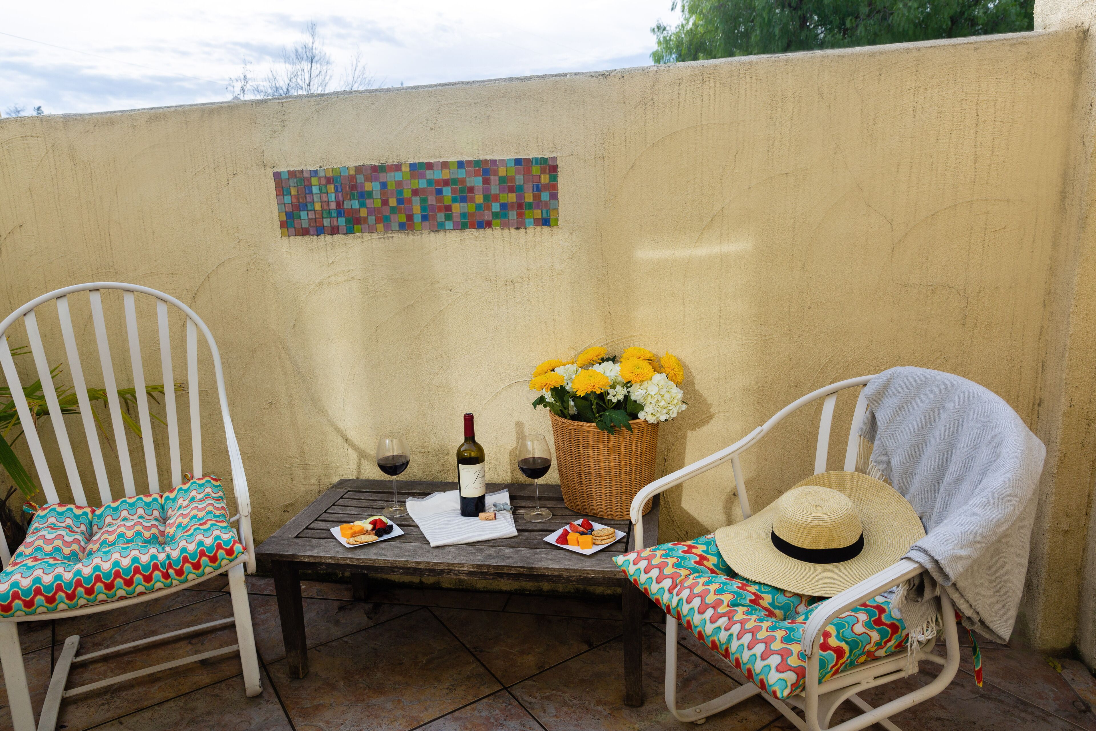 Dos sillas blancas con cojines coloridos, una mesa con una botella de vino, copas y una cesta de flores.