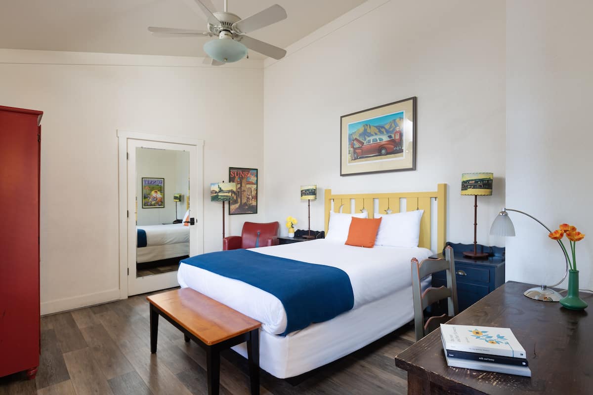 A hotel room with a bed, a desk, a chair, a nightstand, a lamp, a vase with flowers, a ceiling fan, and a painting on the wall.