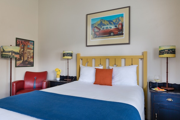 Habitación de hotel con una cama grande, cabecera de madera, una silla roja, una mesita de noche con lámpara y un cuadro en la pared.