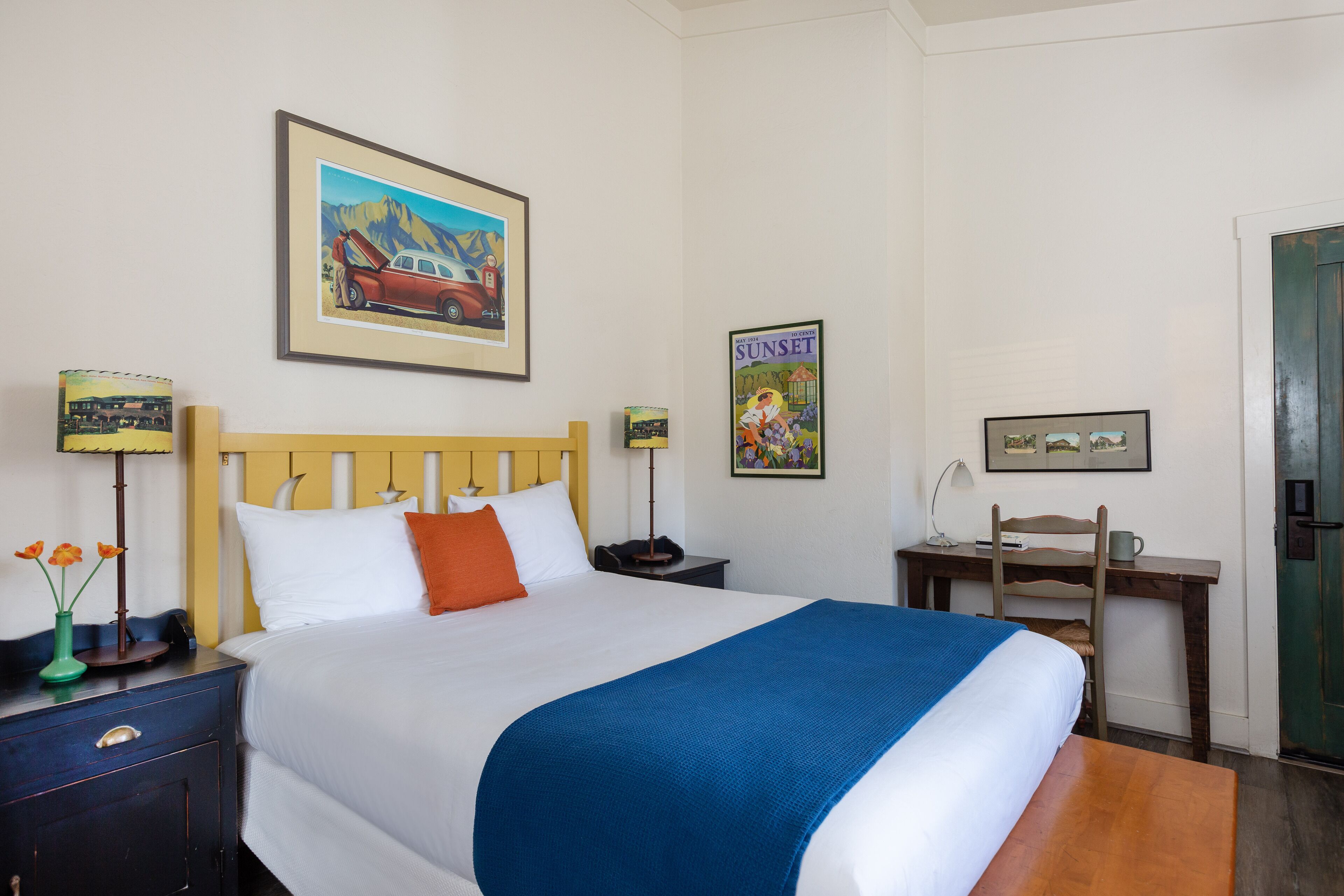 A neatly made bed with a blue blanket, a wooden headboard, and a framed picture of a car on the wall.