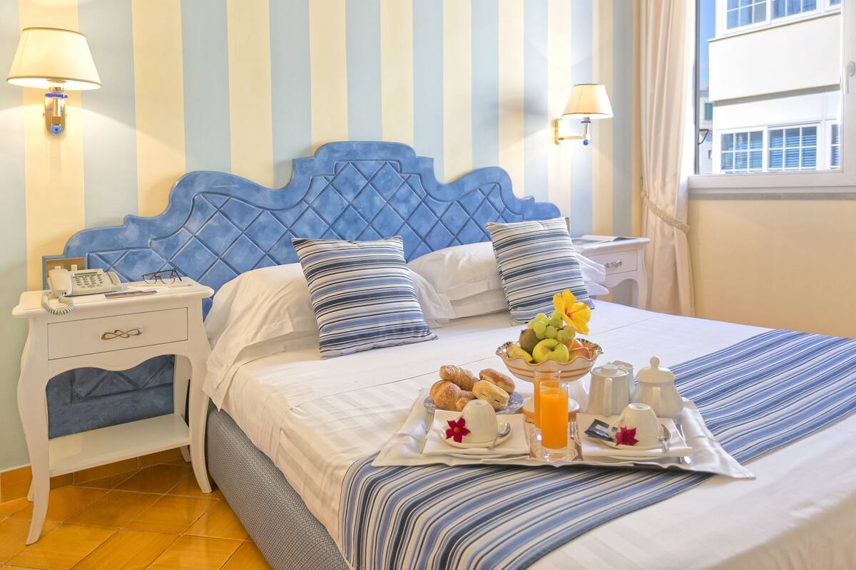 A hotel room with a striped blue and white headboard, a bedside table with a lamp, and a tray of breakfast items including croissants, fruit, and coffee.