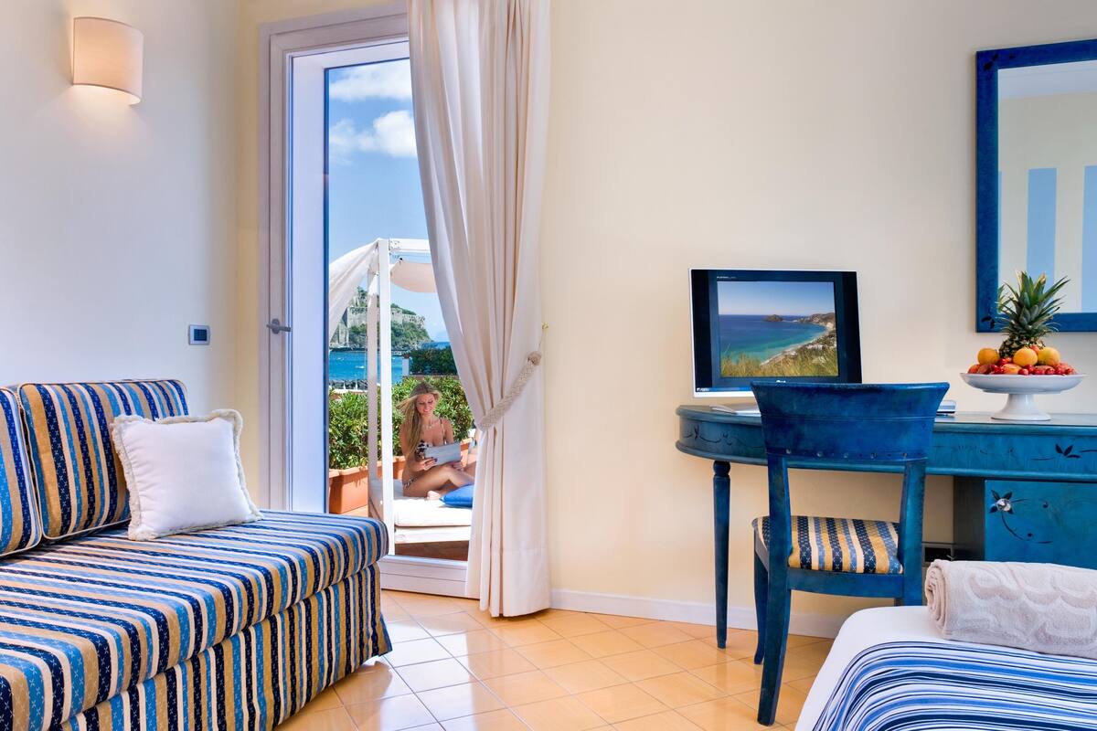 A room with a striped sofa, a desk with a computer, a chair, a bed, and a view of the sea.