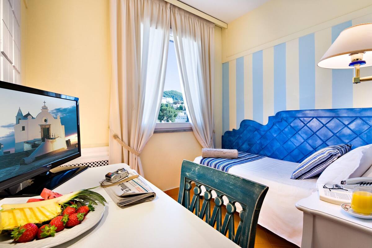 A hotel room with a bed, a desk with a computer, a plate of fruit, and a window with curtains.
