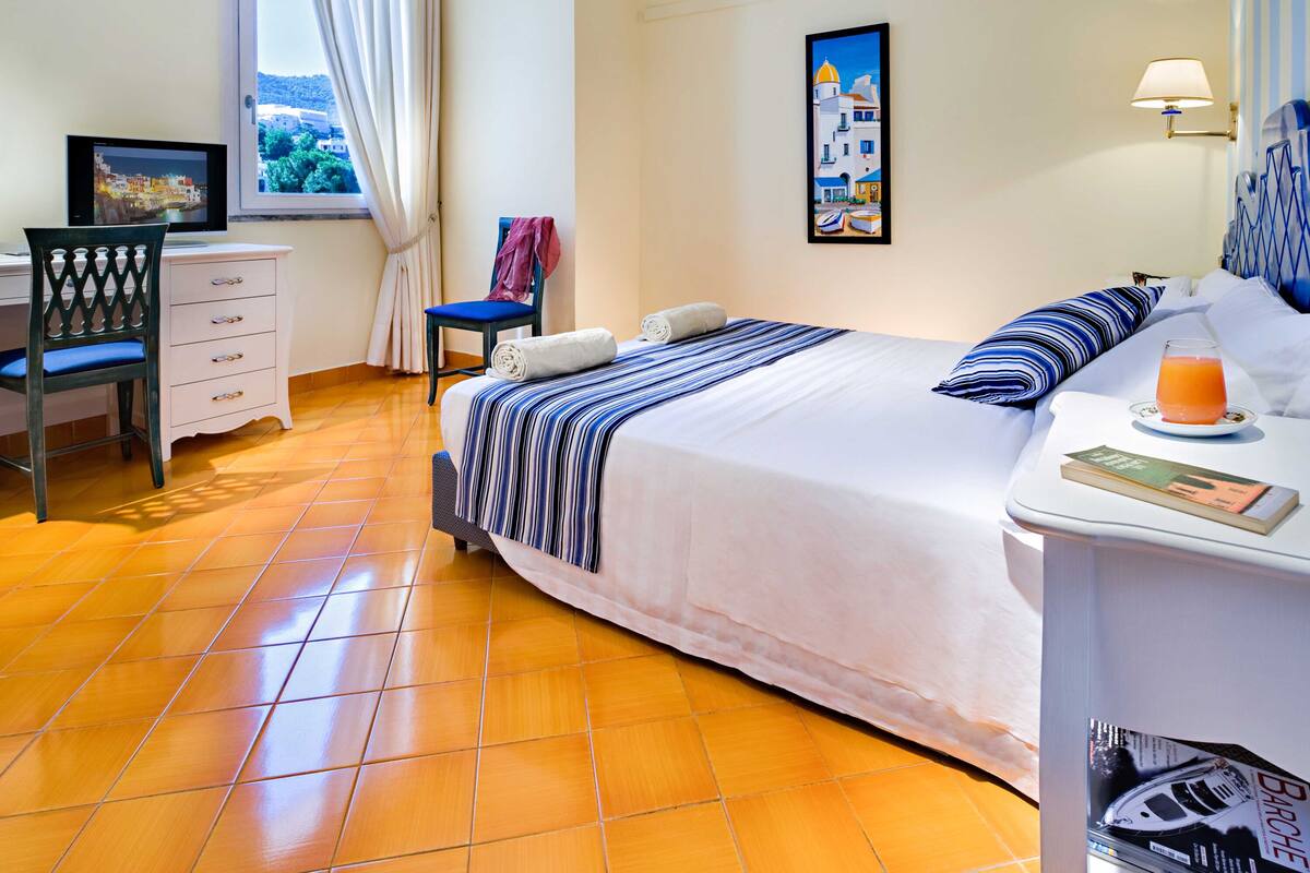 A hotel room with a bed, a desk, a chair, a window with curtains, a wall art, and a bedside table with a book and a glass of orange juice.