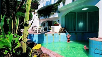 Une piscine avec une personne dans l’eau, entourée de plantes et d’un bâtiment doté d’un balcon incurvé.