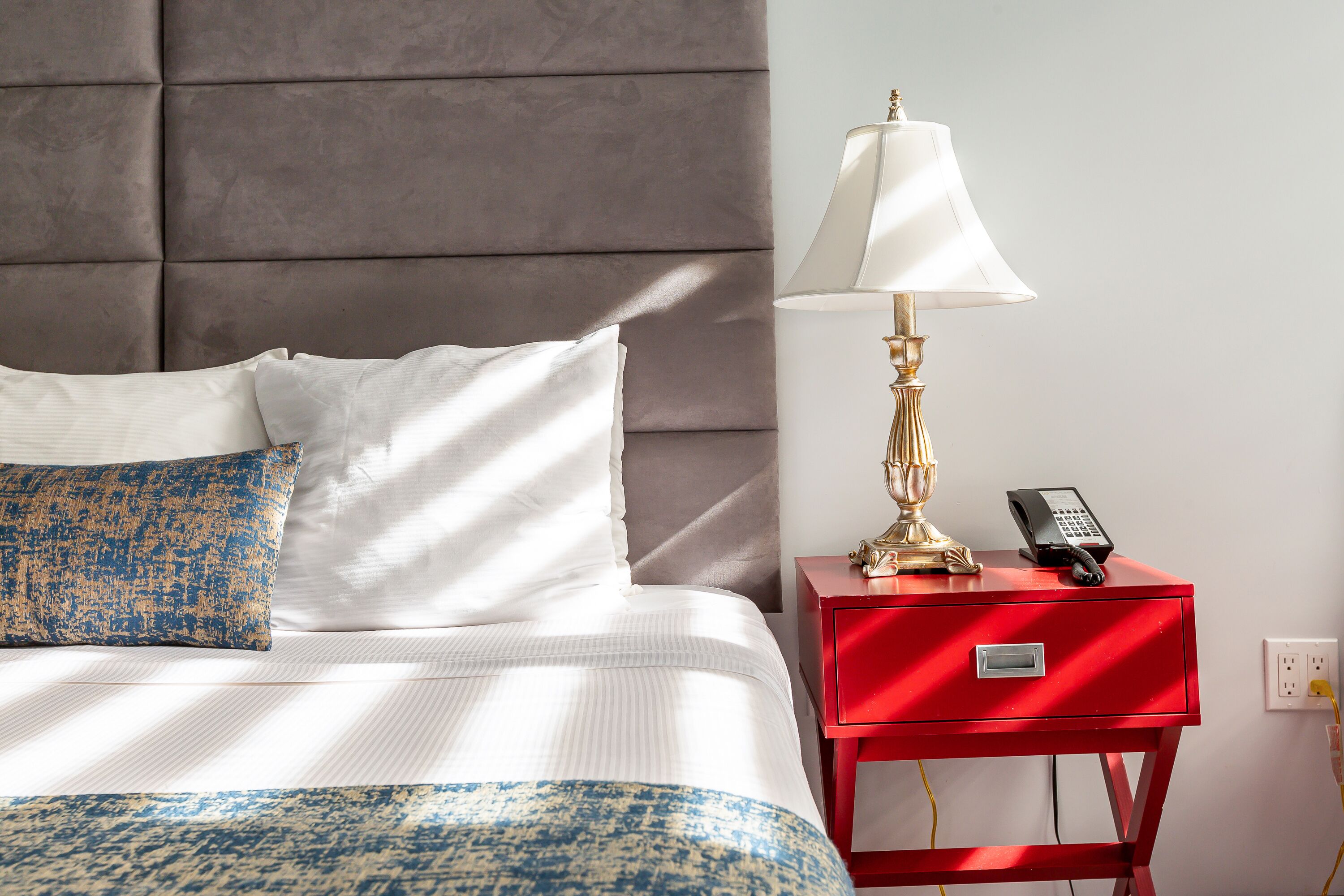 Una cama bien hecha con ropa de cama blanca y azul estampada, una mesita de noche roja con una lámpara y un teléfono, y un cabecero gris.