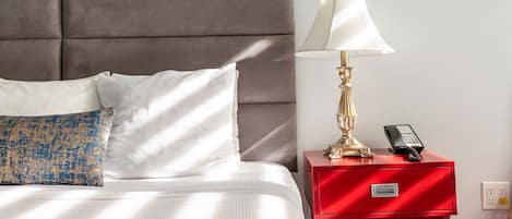 Una cama bien hecha con ropa de cama blanca y azul estampada, una mesita de noche roja con una lámpara y un teléfono, y un cabecero gris.