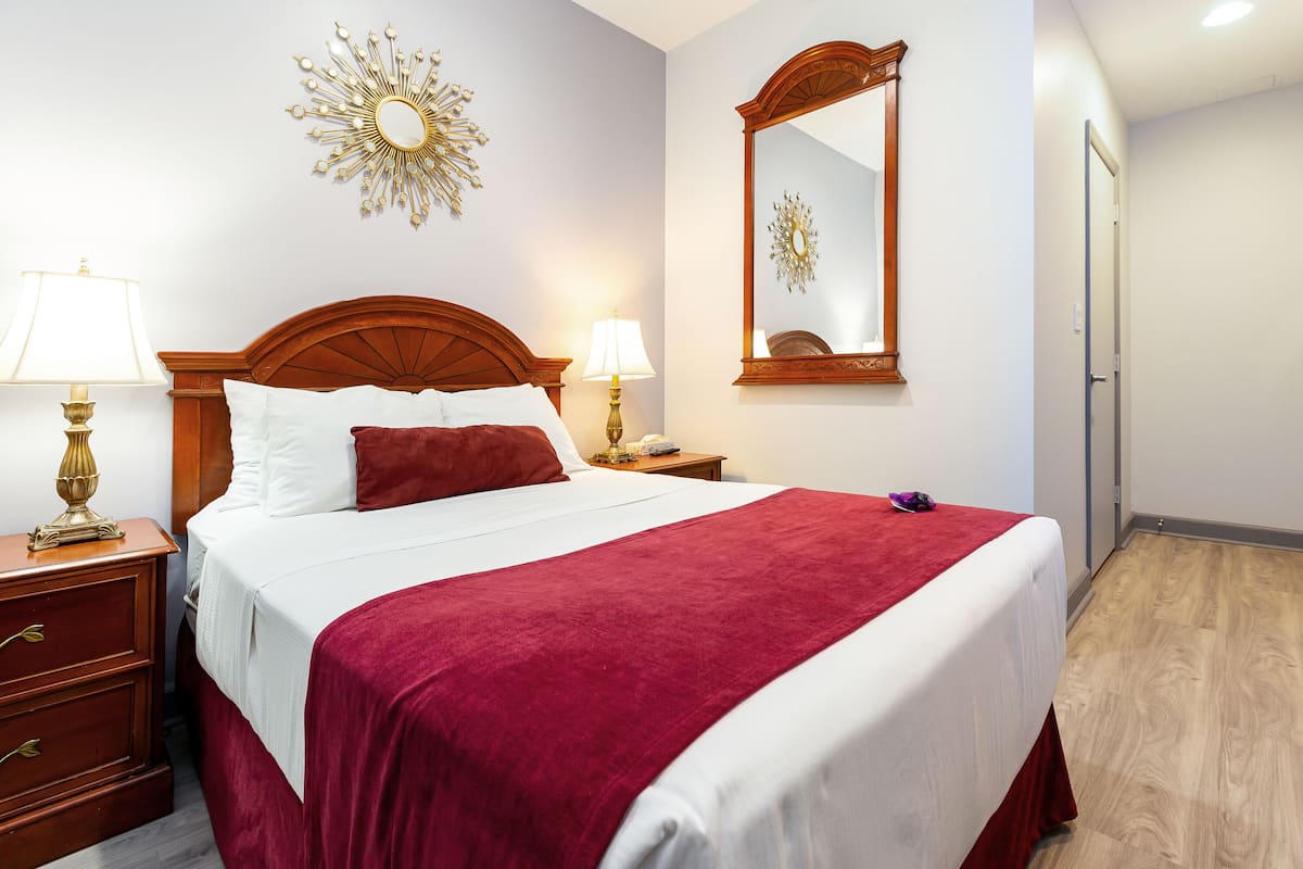 A neatly made bed with a red bedspread, two bedside lamps, a wooden nightstand, and a mirror on the wall.
