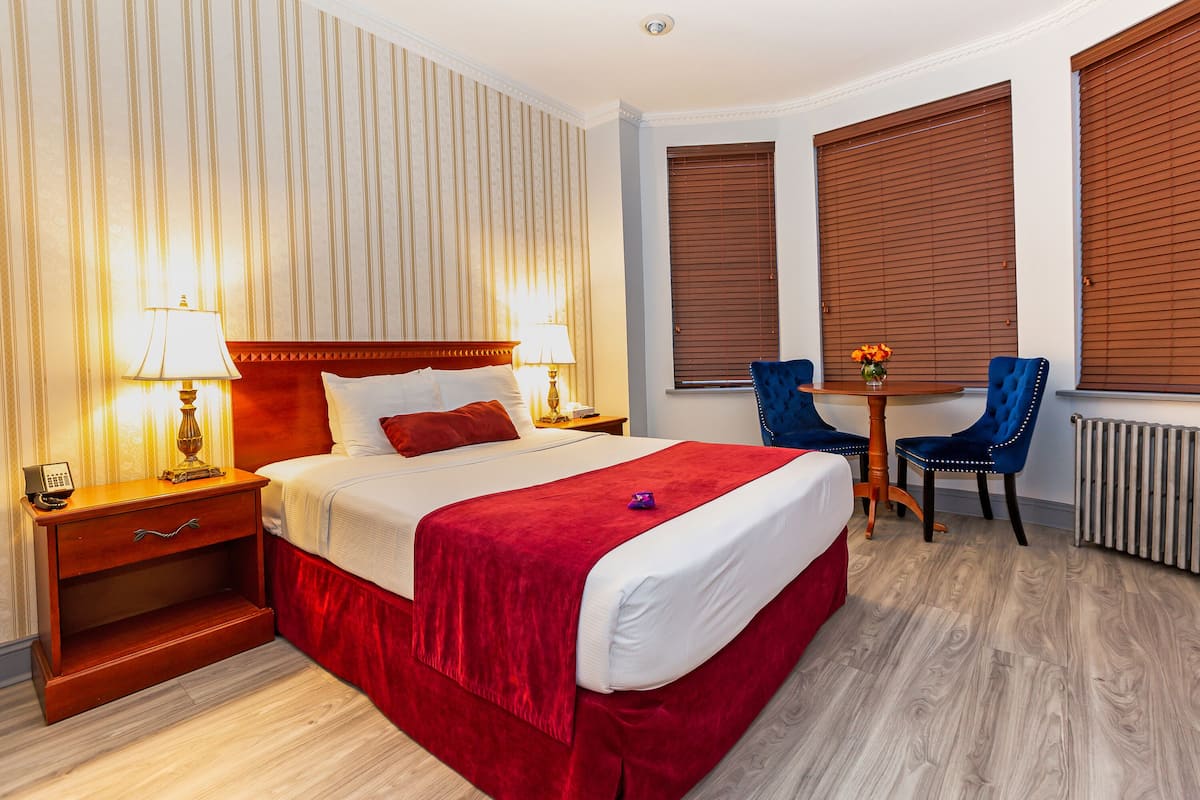 A hotel room with a large bed, a bedside table with a lamp, a small table with chairs, and a window with blinds.