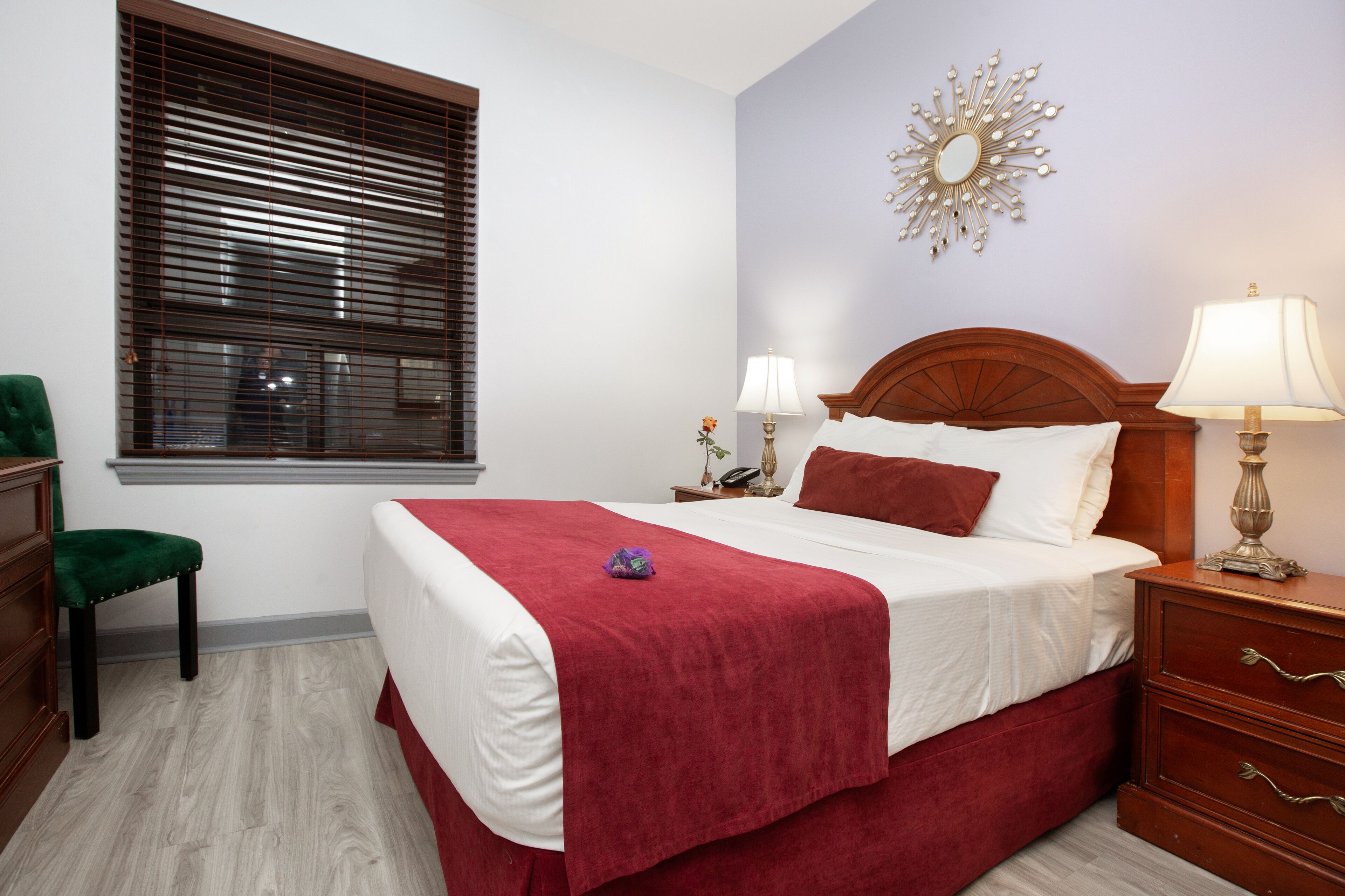 A hotel room with a large bed, a nightstand with a lamp, a green chair, and a window with blinds.