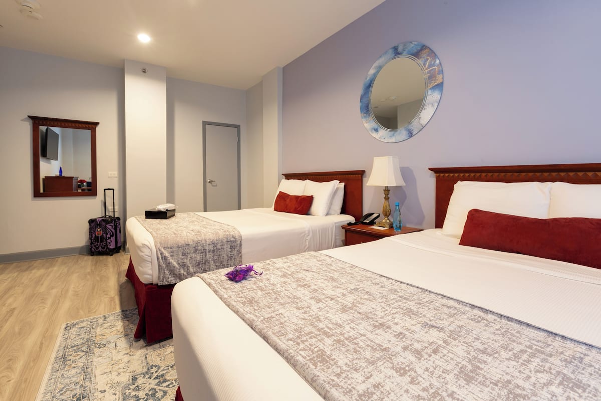 A hotel room with two beds, a wooden floor, a large mirror, and a small table with a lamp.