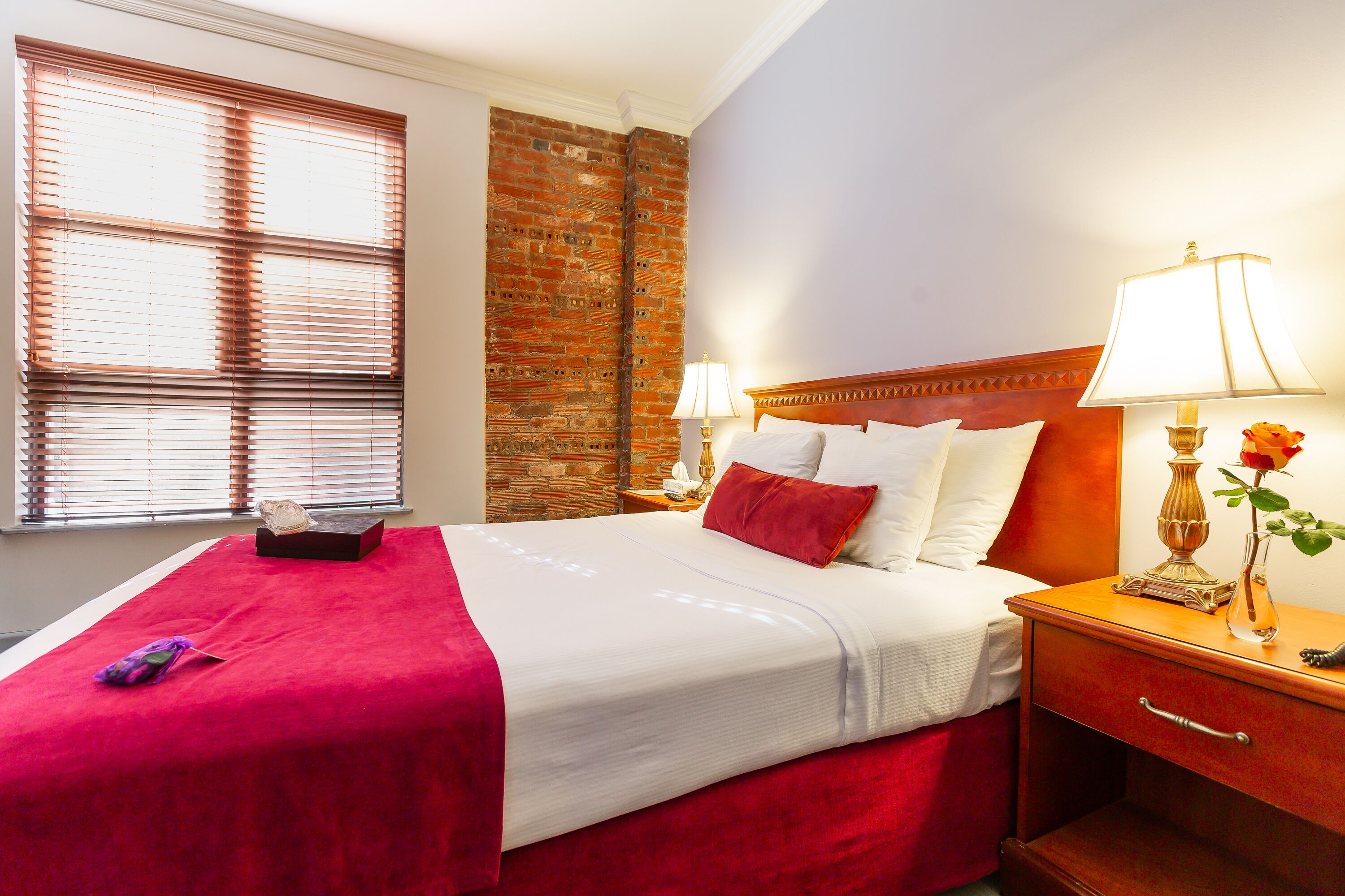 A hotel room with a large bed, a wooden nightstand, a brick wall, and a window with blinds.