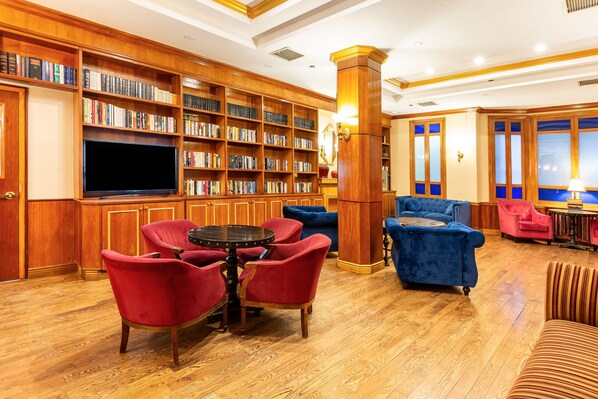 Uma biblioteca com estantes de madeira, uma televisão e poltronas estofadas em vermelho e azul.