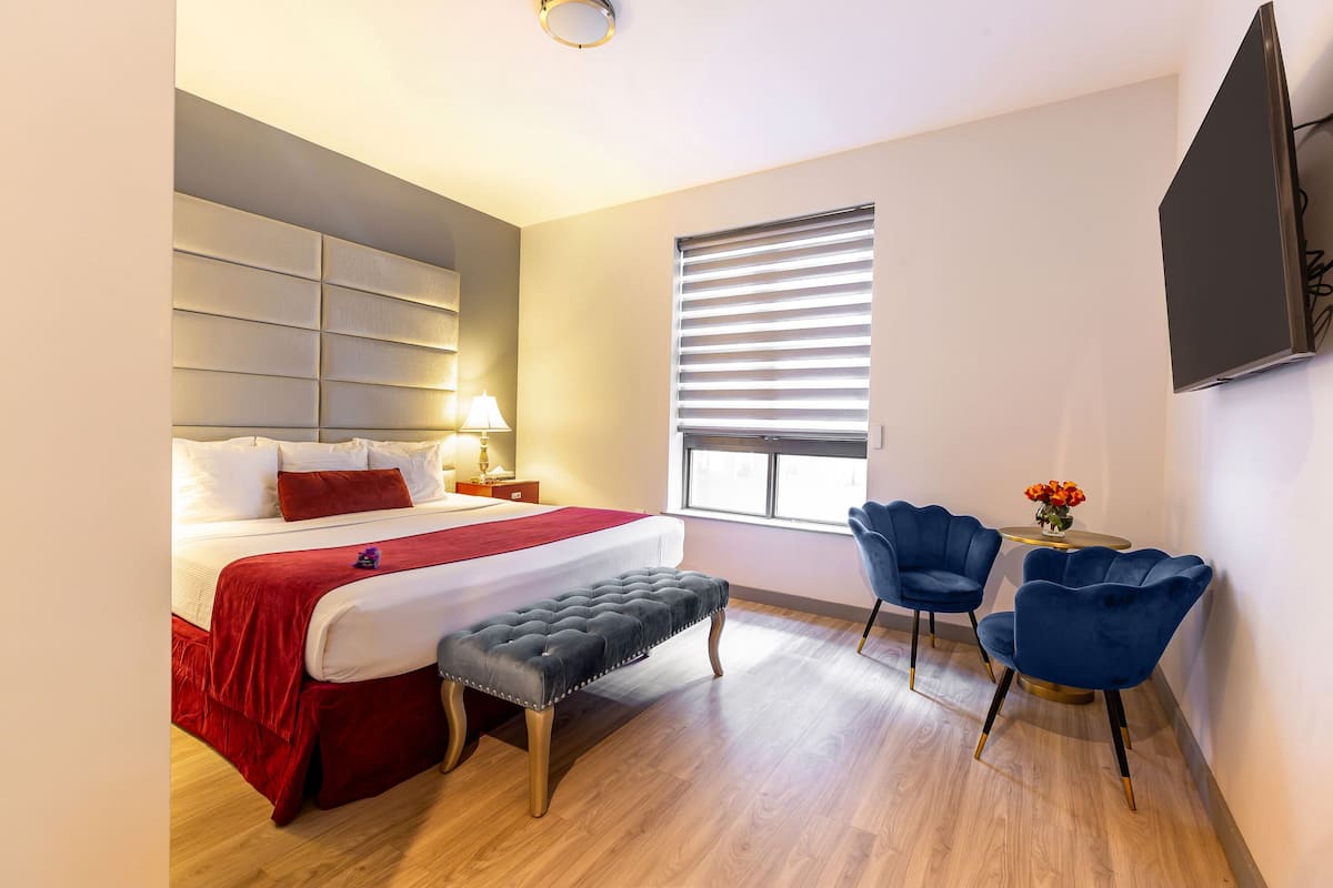A hotel room with a bed, bench, two chairs, a TV, and a window with blinds.
