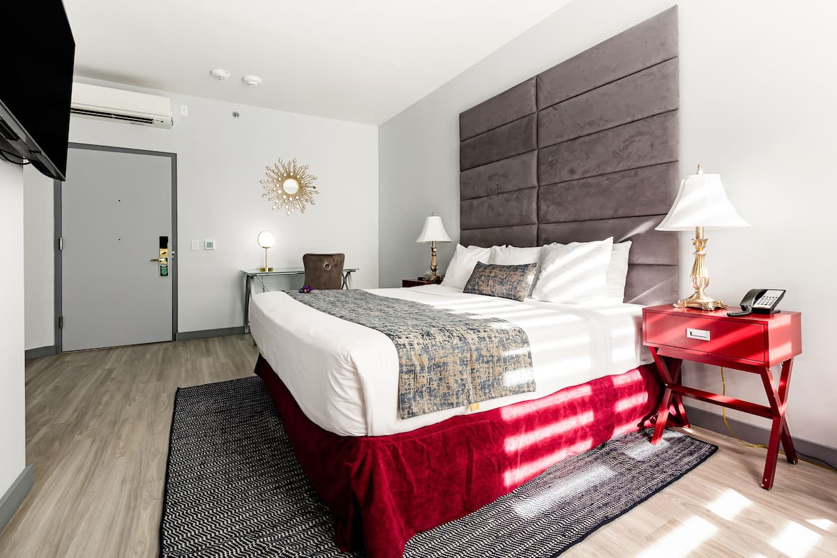 A modern hotel room with a large bed, a red nightstand, a wall-mounted TV, and a decorative sunburst light.