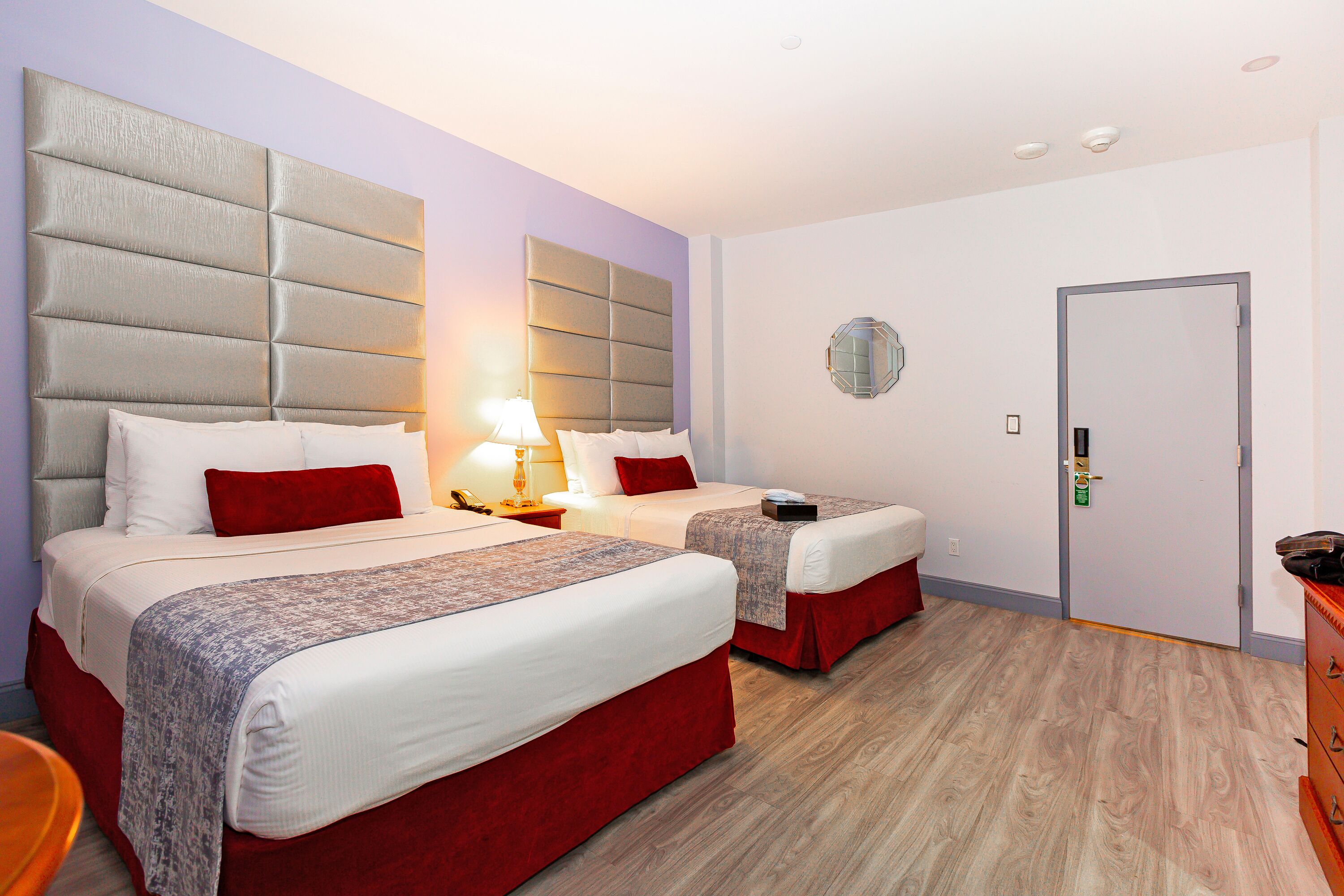 A hotel room with two beds, a bedside table with a lamp, a door, and a dresser.