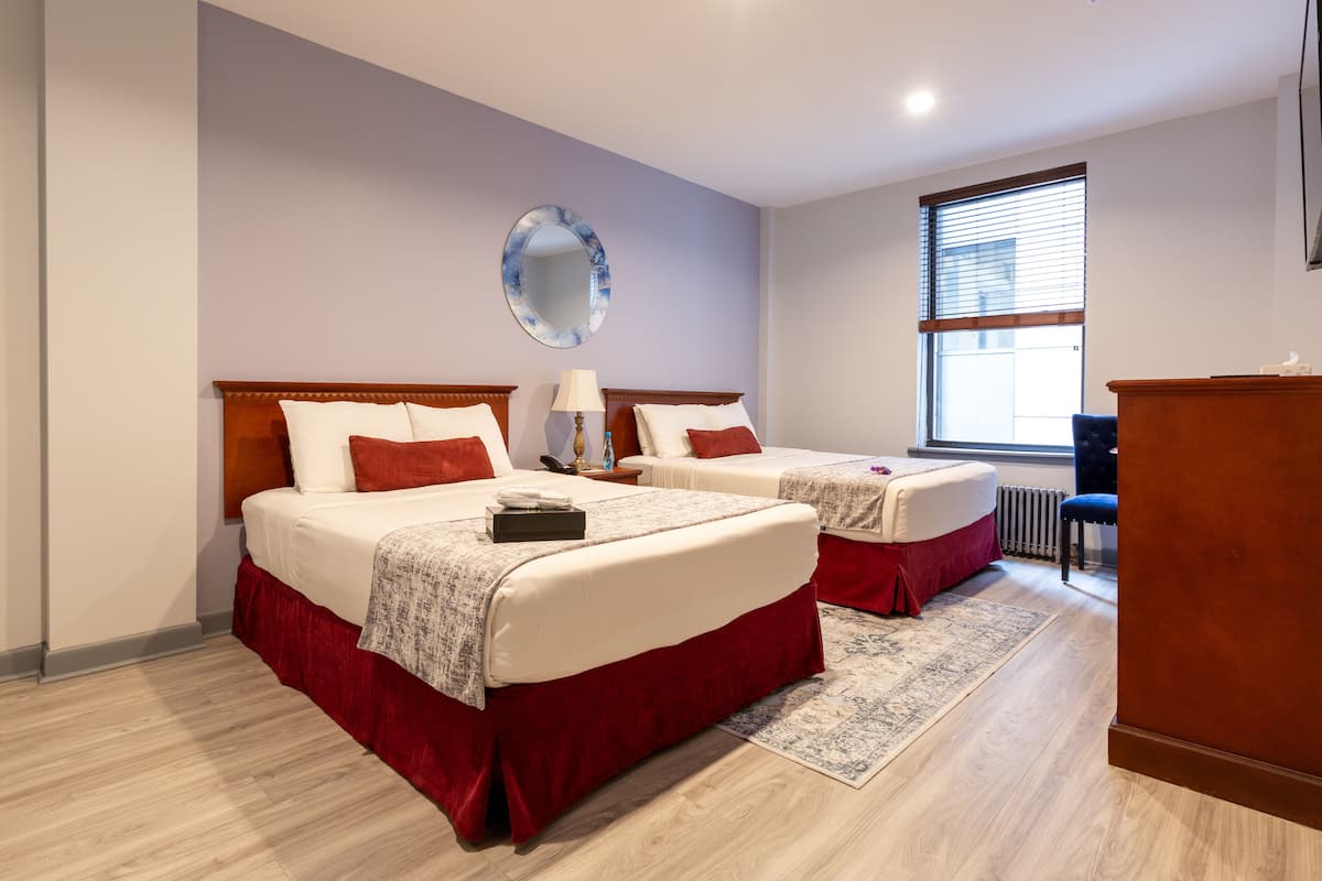 A hotel room with two beds, a large mirror, a window with blinds, and a wooden desk.