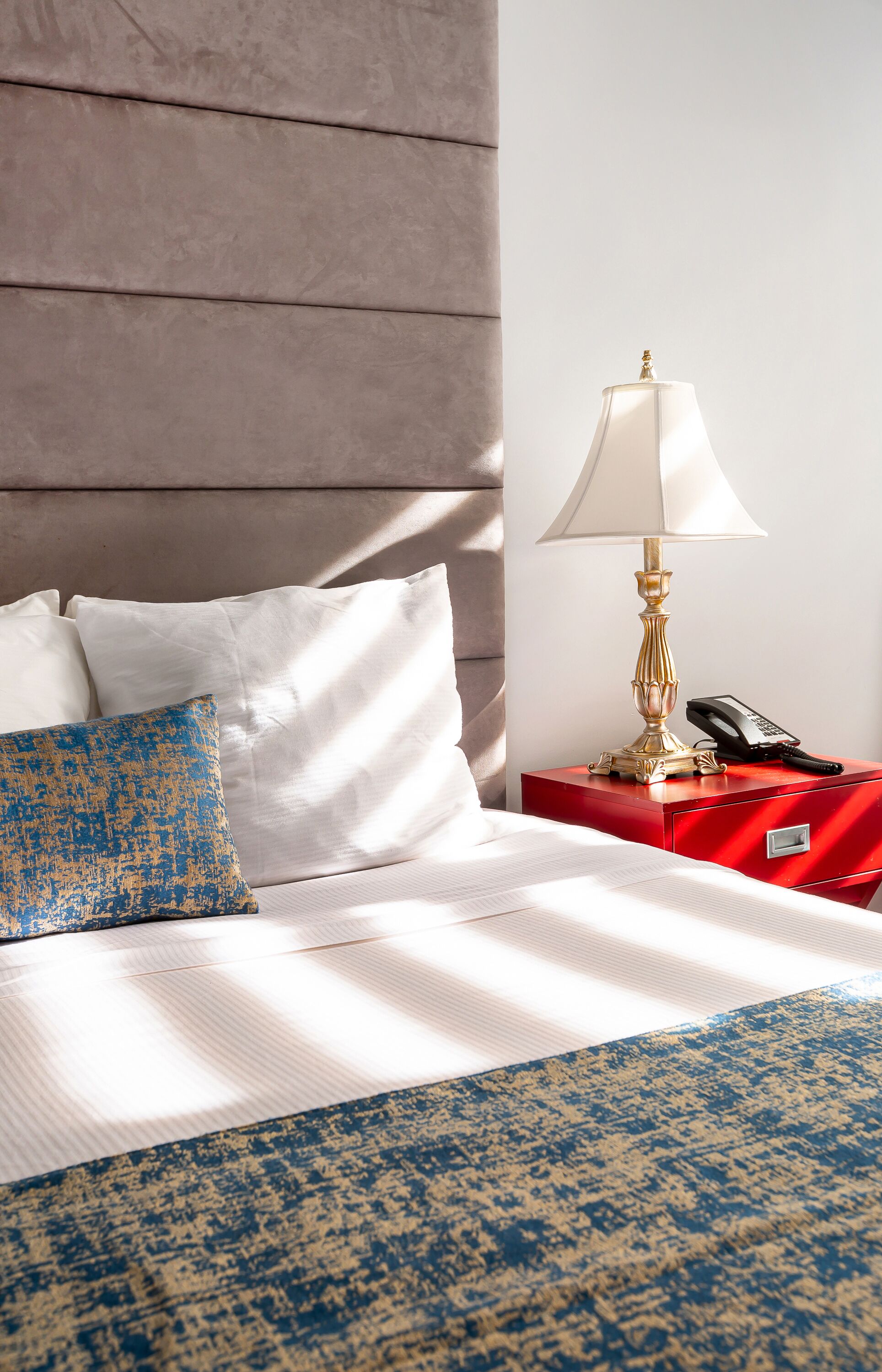 A neatly made bed with white and blue bedding, a red nightstand with a lamp and phone, and a wooden headboard.